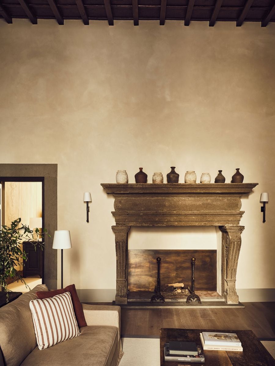A carved stone fireplace with fluted, scrolled corbels anchors a living room at La Réserve Firenze, its mantel lined with a procession of earthenware jars. Clouded plaster walls rise to a dark beamed ceiling, while a stone-framed doorway offers a glimpse of a painted bedroom beyond. A carved stone fireplace with fluted, scrolled corbels anchors a living room at La Réserve Firenze, its mantel lined with a procession of earthenware jars. Clouded plaster walls rise to a dark beamed ceiling, while a stone-framed doorway offers a glimpse of a painted bedroom beyond.