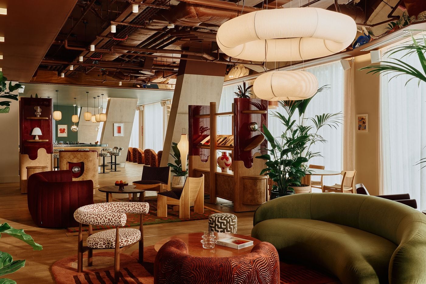 A wide view of the ground-floor lounge reveals Holloway Li’s eclectic take on mid-century modernism, combining olive-green and burgundy seating, graphic textiles, sculptural lighting and glossy bespoke joinery. Exposed ceiling services and angular concrete columns introduce an industrial counterpoint to the space’s softer curves and rich material palette. A wide view of the ground-floor lounge reveals Holloway Li’s eclectic take on mid-century modernism, combining olive-green and burgundy seating, graphic textiles, sculptural lighting and glossy bespoke joinery. Exposed ceiling services and angular concrete columns introduce an industrial counterpoint to the space’s softer curves and rich material palette.