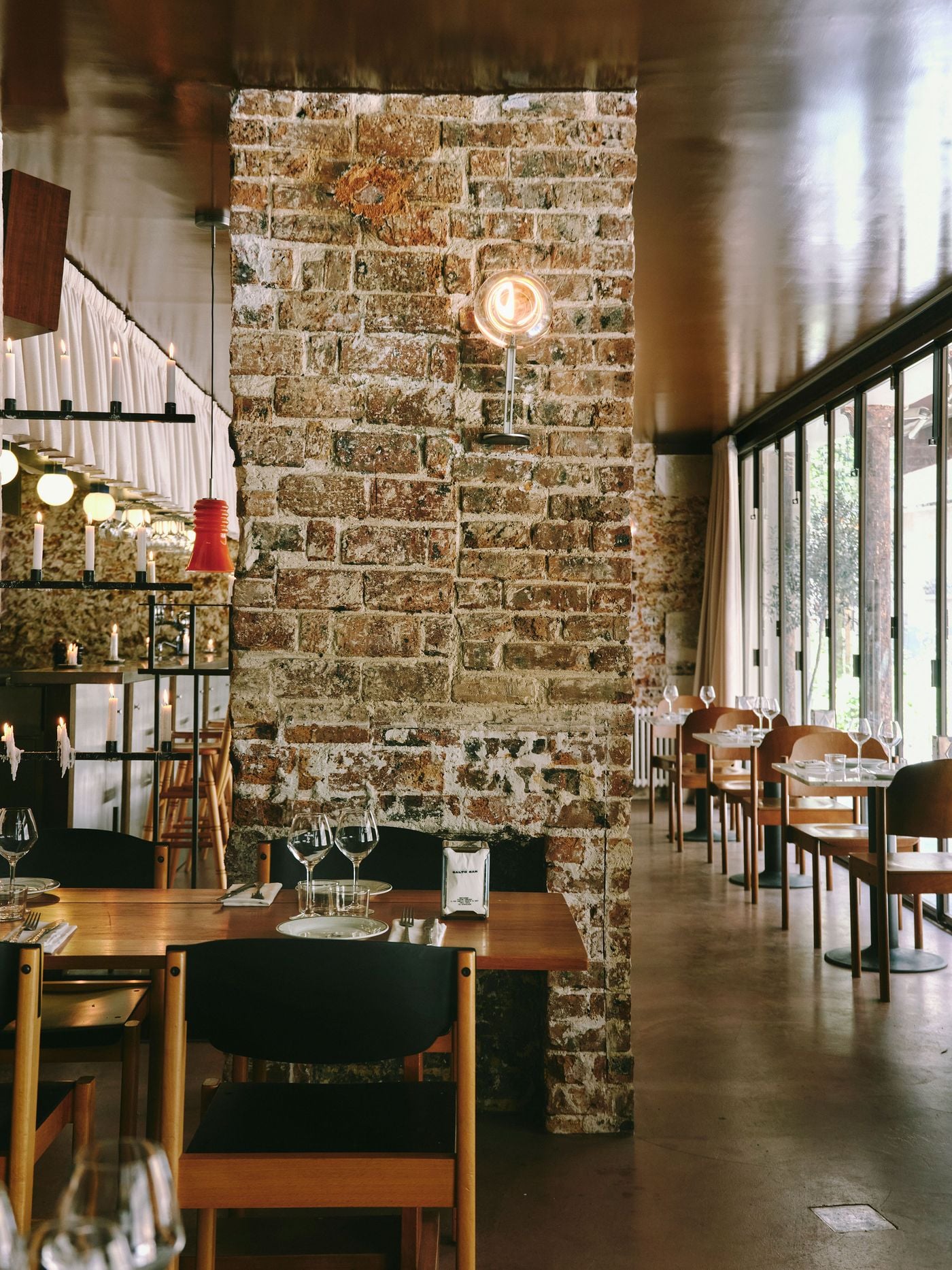 An exposed brick pier, its surface flecked with old plaster, divides the dining room at SaltoBar in Paris. A glass globe sconce glows on the brickwork. To one side, candle-lined metal shelving and a red ribbed pendant frame the bar. To the other, marble tables line a glazed frontage opening onto greenery. An exposed brick pier, its surface flecked with old plaster, divides the dining room at SaltoBar in Paris. A glass globe sconce glows on the brickwork. To one side, candle-lined metal shelving and a red ribbed pendant frame the bar. To the other, marble tables line a glazed frontage opening onto greenery.