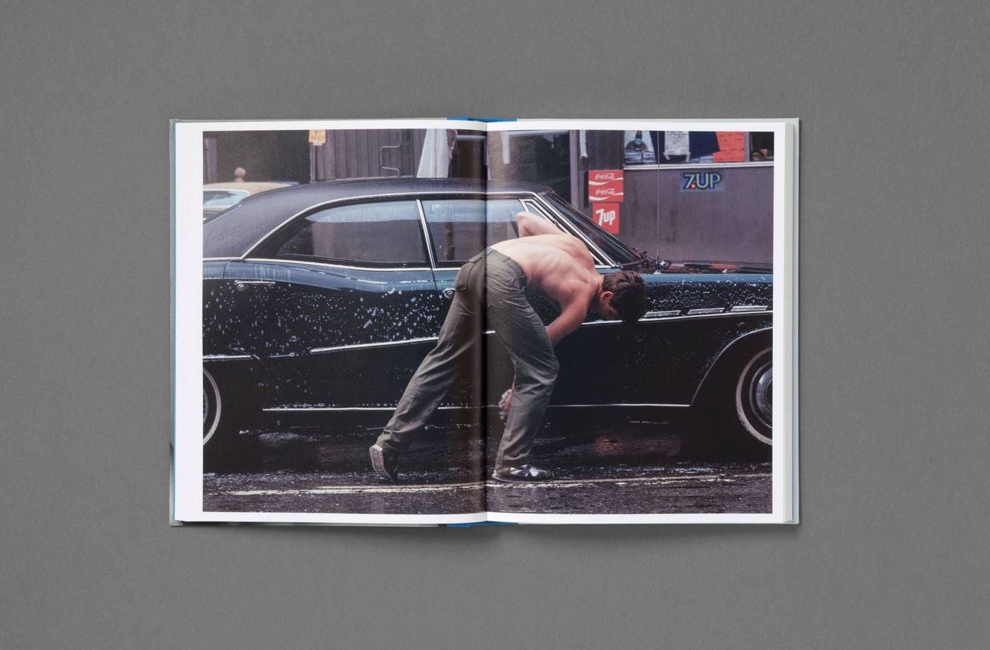 A colour spread from Don't Worry Boys shows a shirtless young man in olive trousers and trainers stooping to wash a gleaming black vintage sedan. Soap suds speckle the dark paintwork, while faded soft-drink signs and a weathered storefront behind him anchor the scene in the street life of 1970s New York. A colour spread from Don't Worry Boys shows a shirtless young man in olive trousers and trainers stooping to wash a gleaming black vintage sedan. Soap suds speckle the dark paintwork, while faded soft-drink signs and a weathered storefront behind him anchor the scene in the street life of 1970s New York.