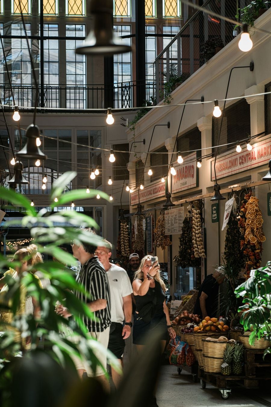 Agora Modiano, Thessaloniki’s restored 1920s covered market: shoppers stroll beneath strings of bulb lights and a steel-and-glass roof, past stalls hung with garlic braids and dried oranges, produce baskets and vintage Central Market signage.