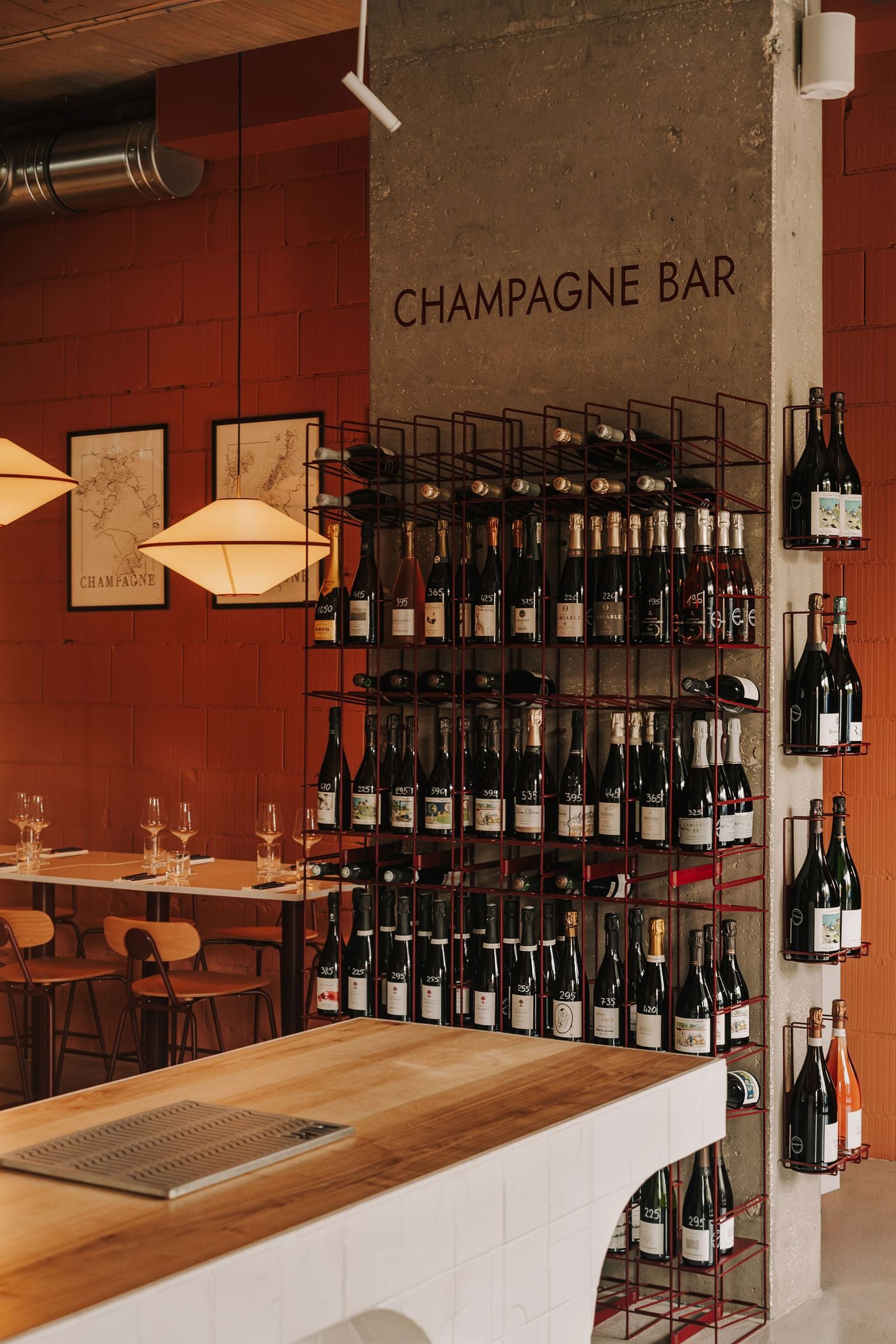 A champagne display at WIN wine bar combines slender burgundy metal shelving with the rough texture of an exposed concrete column. Bottles arranged vertically and horizontally create a dense geometric composition, while sculptural pendant lights illuminate an intimate seating area framed by terracotta-toned masonry walls. A champagne display at WIN wine bar combines slender burgundy metal shelving with the rough texture of an exposed concrete column. Bottles arranged vertically and horizontally create a dense geometric composition, while sculptural pendant lights illuminate an intimate seating area framed by terracotta-toned masonry walls.