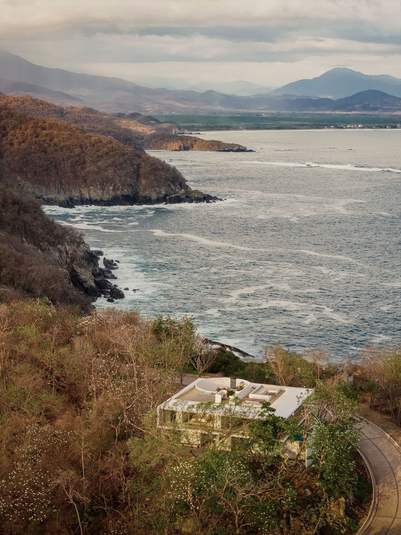 An aerial view of Casa Malonic by HW Studio in Zihuatanejo, Mexico, shows the white modernist volume set among sun-browned woodland flecked with white blossoms above the Pacific. The circular courtyard and rooftop lounge read clearly from above, while a curving road skirts the house and rocky headlands recede towards distant, mist-veiled mountains. An aerial view of Casa Malonic by HW Studio in Zihuatanejo, Mexico, shows the white modernist volume set among sun-browned woodland flecked with white blossoms above the Pacific. The circular courtyard and rooftop lounge read clearly from above, while a curving road skirts the house and rocky headlands recede towards distant, mist-veiled mountains.