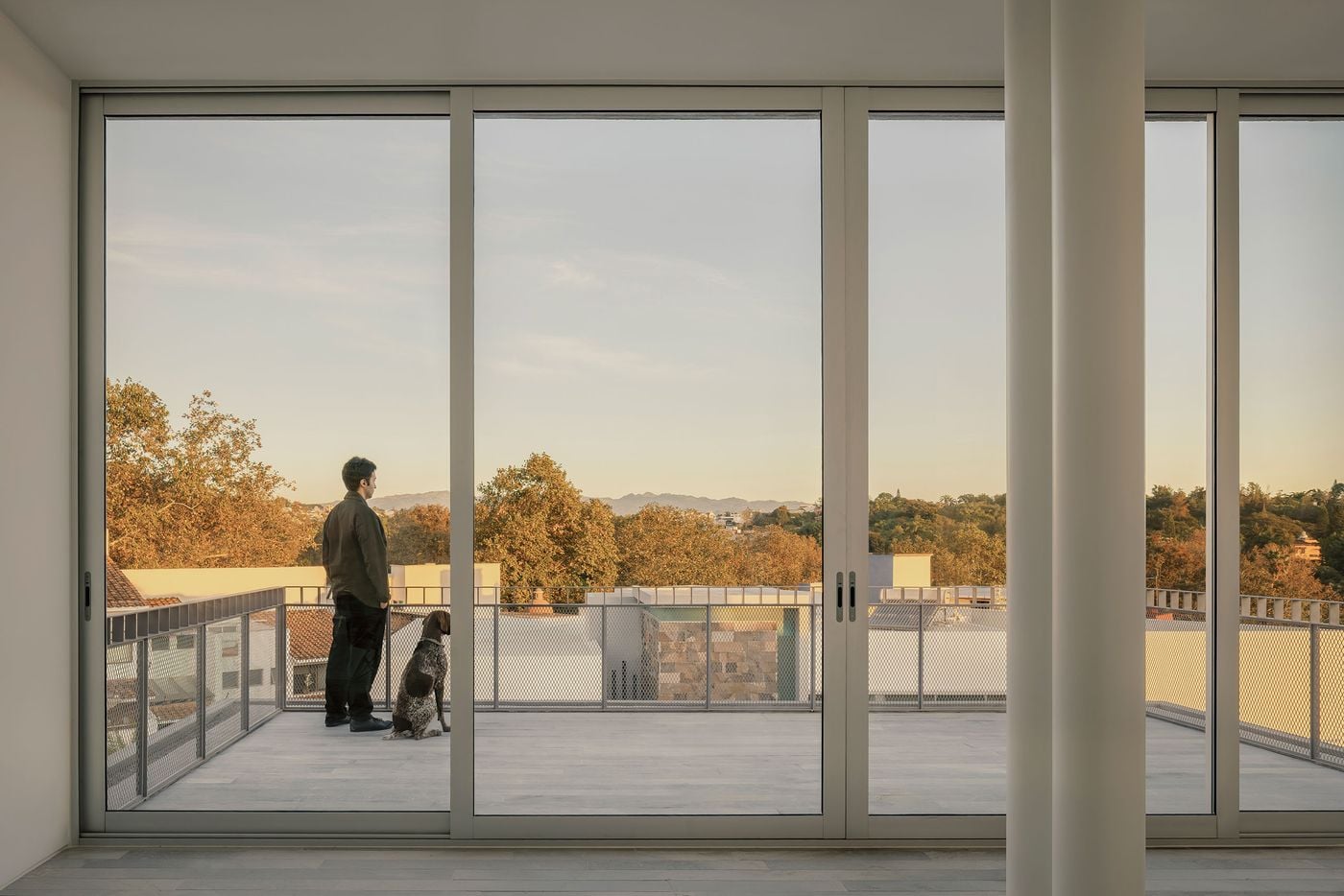 Full-height sliding glass doors frame Casa Tlaloc’s upper terrace like a panoramic window, extending the minimalist interior towards treetops and distant mountains. An occupant and dog stand quietly at the metal-mesh balustrade in the warm evening light, emphasising the house’s focus on observation, stillness and connection to its surroundings. Full-height sliding glass doors frame Casa Tlaloc’s upper terrace like a panoramic window, extending the minimalist interior towards treetops and distant mountains. An occupant and dog stand quietly at the metal-mesh balustrade in the warm evening light, emphasising the house’s focus on observation, stillness and connection to its surroundings.