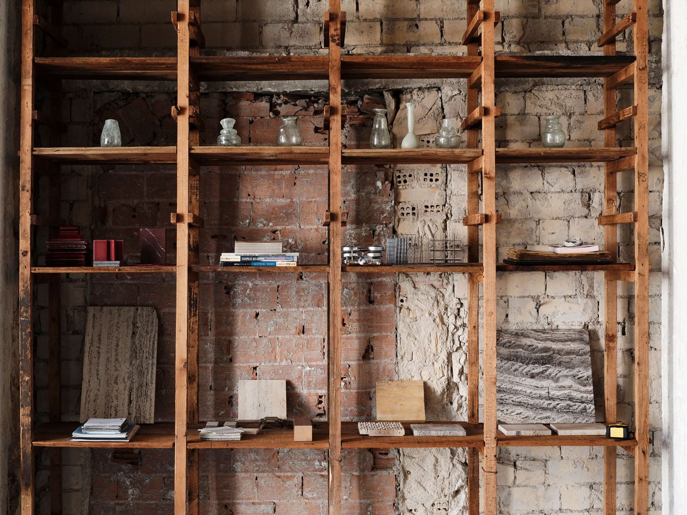 A floor-to-ceiling timber shelving system displays material samples, books, papers and glass vessels against a patchwork wall of exposed brick and masonry. Its warm, weathered wood complements the imperfect historic surfaces, turning a utilitarian storage element into a showcase of Castor’s working material library. A floor-to-ceiling timber shelving system displays material samples, books, papers and glass vessels against a patchwork wall of exposed brick and masonry. Its warm, weathered wood complements the imperfect historic surfaces, turning a utilitarian storage element into a showcase of Castor’s working material library.