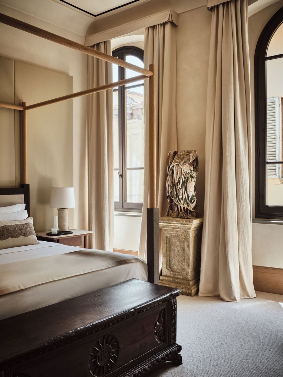 A weathered polychrome figure, its painted drapery flaking to reveal the material beneath, stands on a carved stone pedestal between linen-curtained arched windows in a bedroom at La Réserve Firenze. In the foreground, an antique chest carved with a sunburst emblem anchors a pale oak four-poster bed. A weathered polychrome figure, its painted drapery flaking to reveal the material beneath, stands on a carved stone pedestal between linen-curtained arched windows in a bedroom at La Réserve Firenze. In the foreground, an antique chest carved with a sunburst emblem anchors a pale oak four-poster bed.