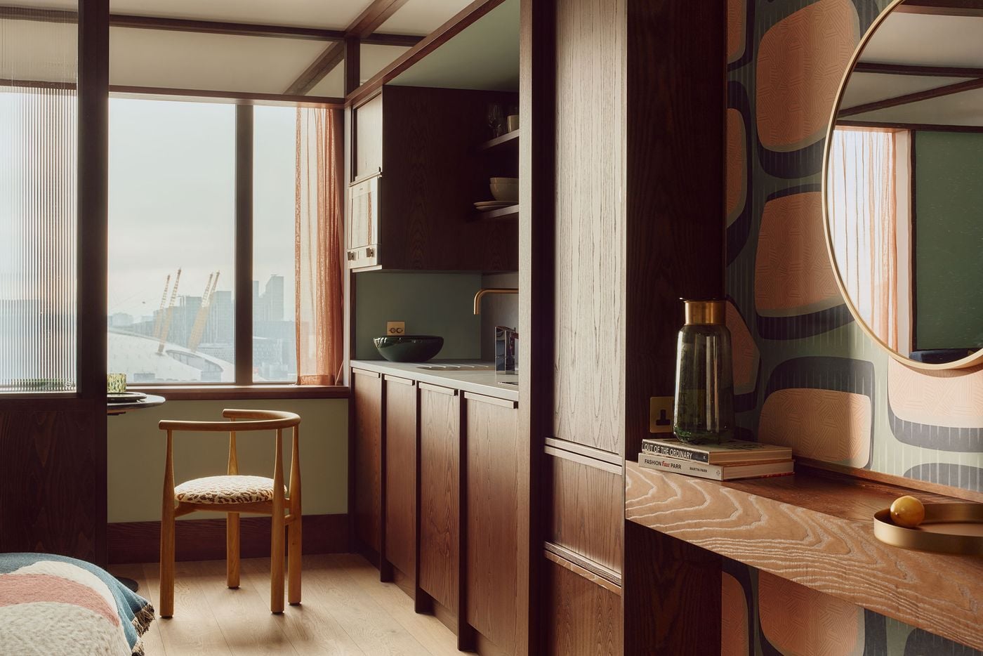 Warm walnut cabinetry integrates the guest room’s kitchenette and dressing area, while a patterned wall panel and circular mirror add a graphic mid-century note. Beyond, a dining chair faces expansive windows overlooking the Thames, balancing compact apartment-style functionality with an unexpectedly generous sense of outlook. Warm walnut cabinetry integrates the guest room’s kitchenette and dressing area, while a patterned wall panel and circular mirror add a graphic mid-century note. Beyond, a dining chair faces expansive windows overlooking the Thames, balancing compact apartment-style functionality with an unexpectedly generous sense of outlook.
