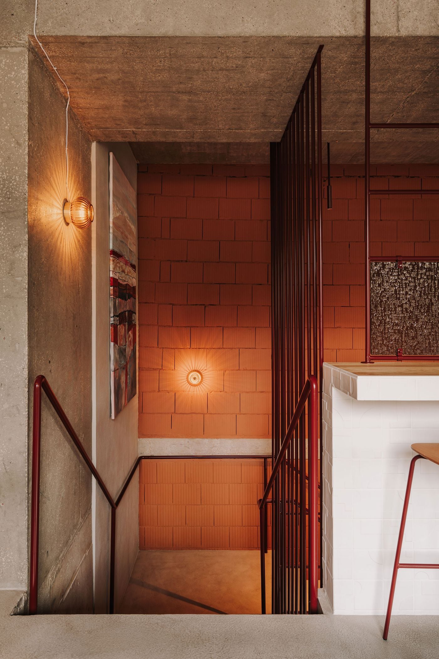 The staircase connecting WIN wine bar’s two levels is framed by slender burgundy metal railings, raw concrete and terracotta-coloured masonry. Ribbed glass wall sconces cast radiating patterns of warm light across the rugged surfaces, while an abstract painting introduces additional colour and texture to the restrained industrial composition. The staircase connecting WIN wine bar’s two levels is framed by slender burgundy metal railings, raw concrete and terracotta-coloured masonry. Ribbed glass wall sconces cast radiating patterns of warm light across the rugged surfaces, while an abstract painting introduces additional colour and texture to the restrained industrial composition.