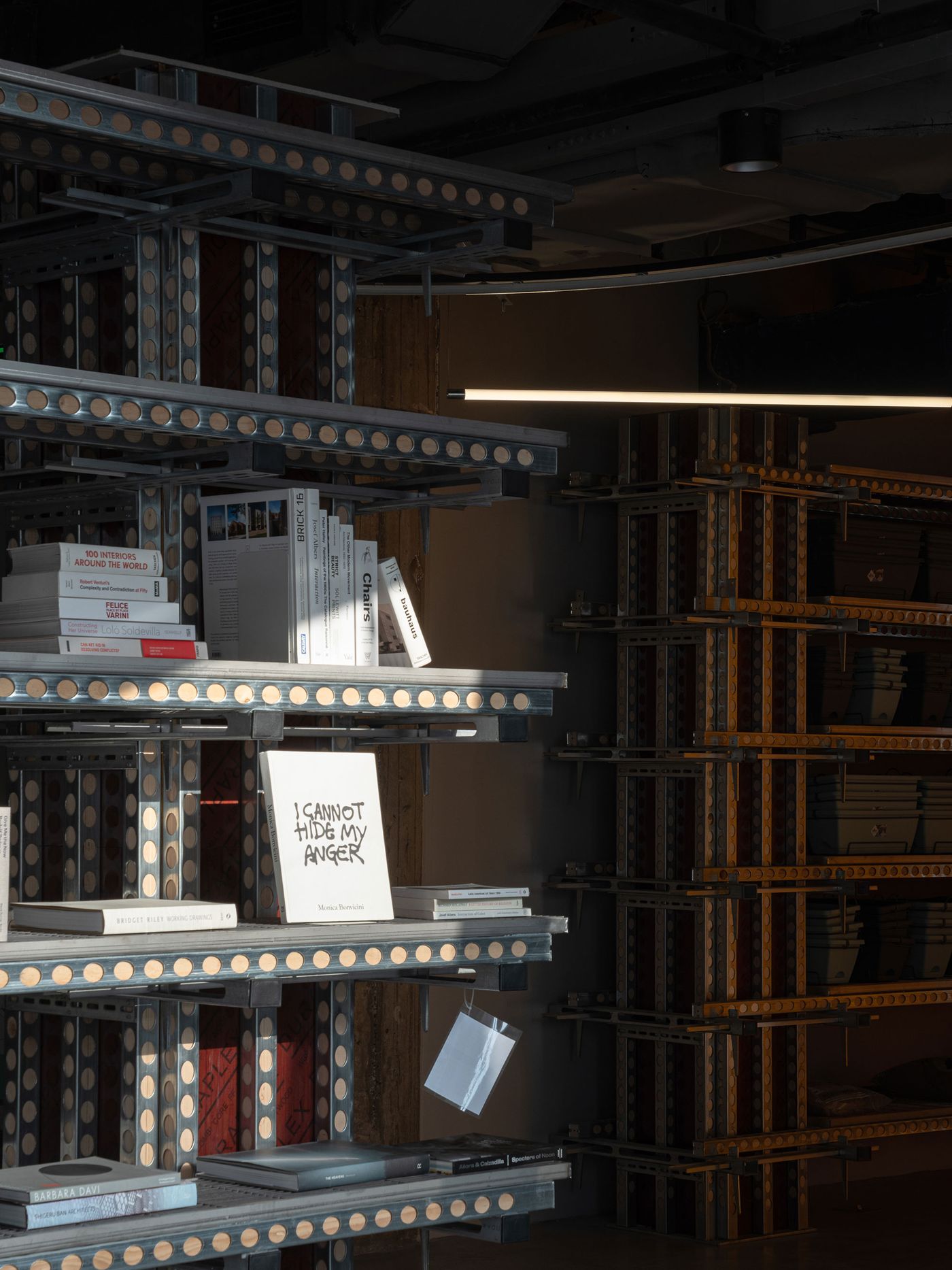 A striking close-up of the multi-tiered, modular shelving system in 889 GLO Art Space, crafted from perforated metal and dark, textured panels. The shelves are thoughtfully arranged with a curated selection of art books, including one prominently titled "I CANNOT HIDE MY ANGER." A single horizontal light strip illuminates the industrial structure, highlighting the space's commitment to both brutalist aesthetics and intellectual curation.