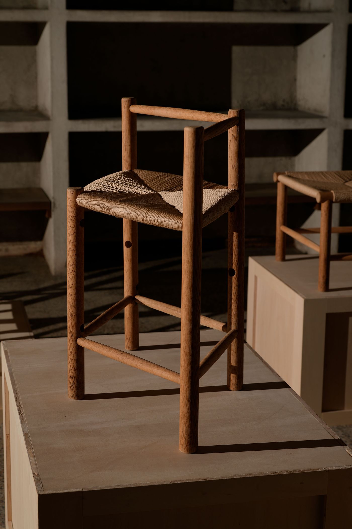 A wooden chair with a woven seat stands on a light-colored pedestal in a well-lit space, emphasizing its minimalist design. A wooden chair with a woven seat stands on a light-colored pedestal in a well-lit space, emphasizing its minimalist design.