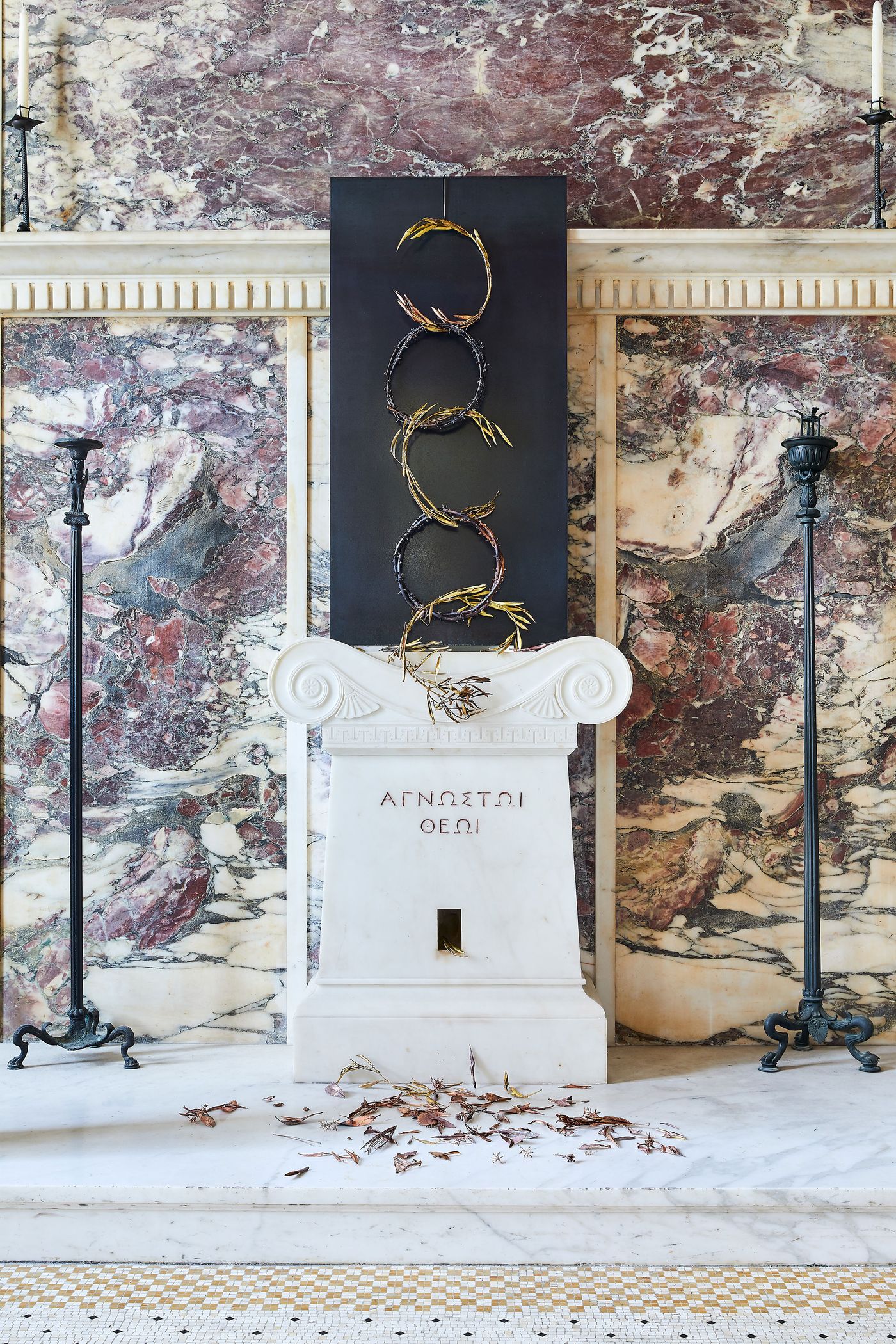 A modern art installation by Gabriel Leger is set against a black panel, mounted above a white marble altar. The sculpture features interwoven, circular wreaths of gilded, thorned branches. Below, metallic leaves are scattered across the floor, referencing the classical inscription on the altar, which reads "To the Unknown God."