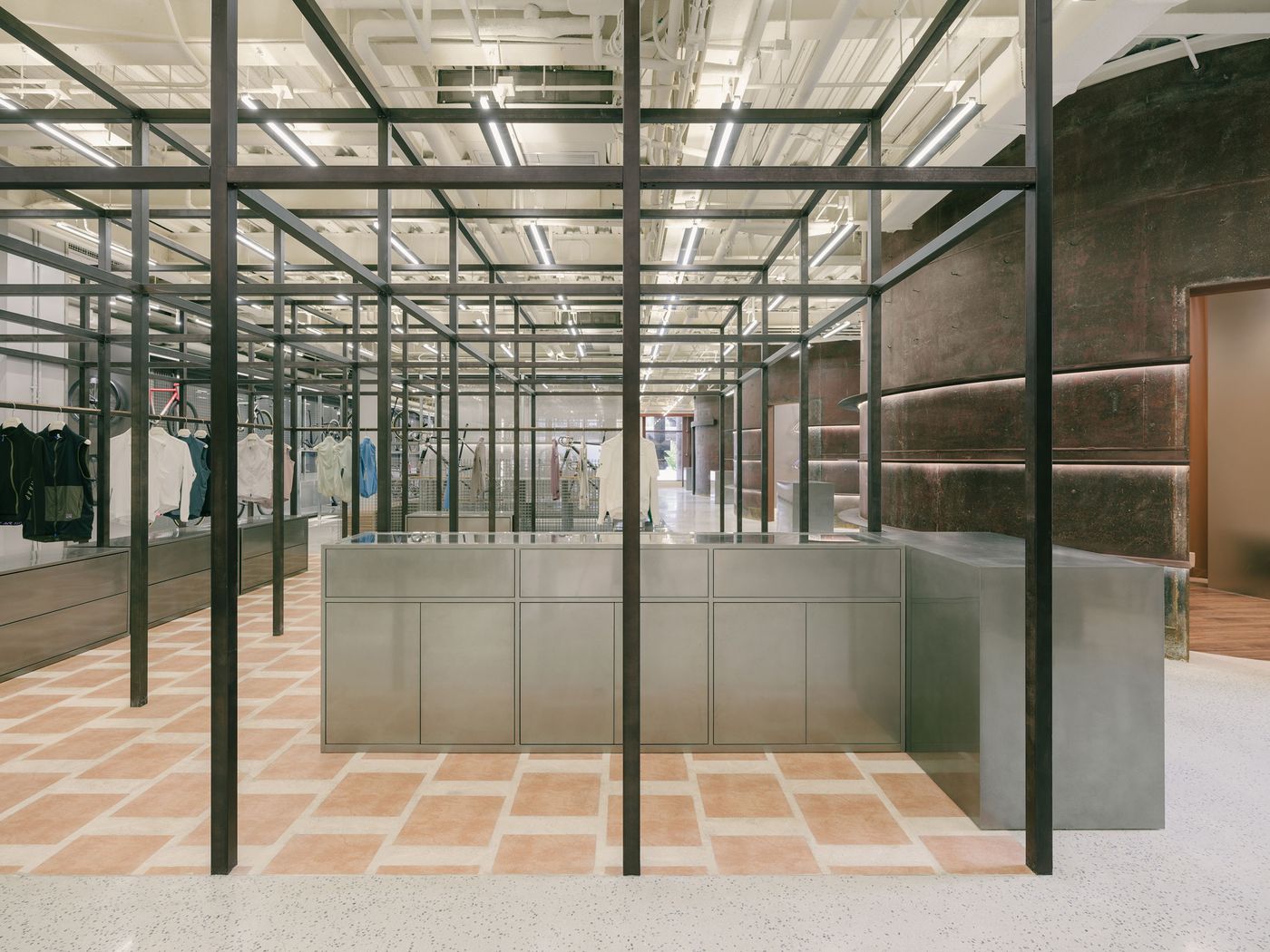A wide-angle interior view of AITASHOP showcases the extensive, linear retail layout defined by a commanding grid of black metal framework that delineates display areas for apparel on sleek, stainless steel counters. The subtle shift from polished concrete to terracotta-tiled flooring visually guides visitors through the sophisticated, industrial-inspired space, illuminated by overhead linear lighting. A wide-angle interior view of AITASHOP showcases the extensive, linear retail layout defined by a commanding grid of black metal framework that delineates display areas for apparel on sleek, stainless steel counters. The subtle shift from polished concrete to terracotta-tiled flooring visually guides visitors through the sophisticated, industrial-inspired space, illuminated by overhead linear lighting.