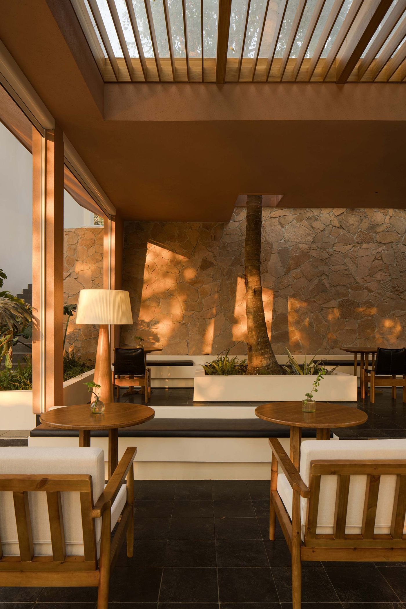 Modern indoor seating area with wooden furniture, a tall lamp, and stone walls illuminated by warm sunlight filtering through slatted skylights. Modern indoor seating area with wooden furniture, a tall lamp, and stone walls illuminated by warm sunlight filtering through slatted skylights.