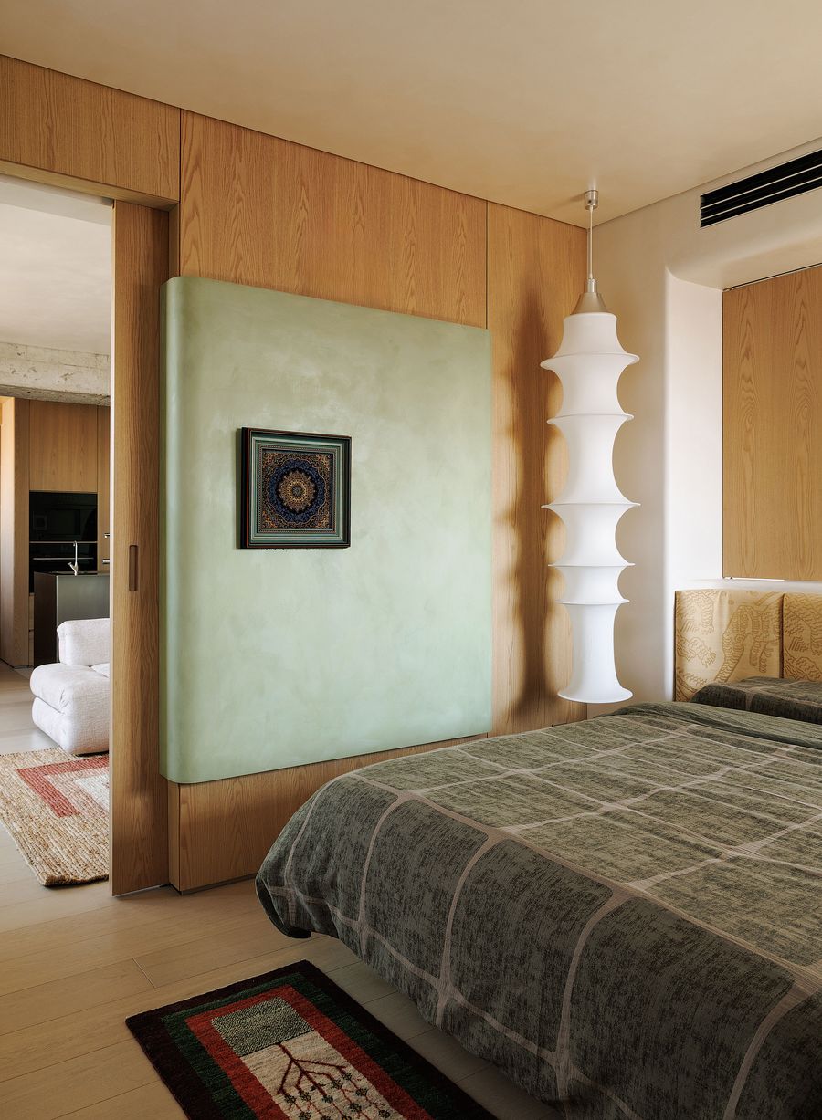 A tranquil bedroom space featuring a unique, tiered sculptural pendant light that adds a dramatic artistic element. The room is defined by warm wood paneling and a soft sage green headboard wall adorned with a framed, intricately detailed artwork. A traditional patterned rug lies on the light wood floor, leading to an adjacent living area, suggesting a thoughtful blend of modern and classic aesthetics. A tranquil bedroom space featuring a unique, tiered sculptural pendant light that adds a dramatic artistic element. The room is defined by warm wood paneling and a soft sage green headboard wall adorned with a framed, intricately detailed artwork. A traditional patterned rug lies on the light wood floor, leading to an adjacent living area, suggesting a thoughtful blend of modern and classic aesthetics.
