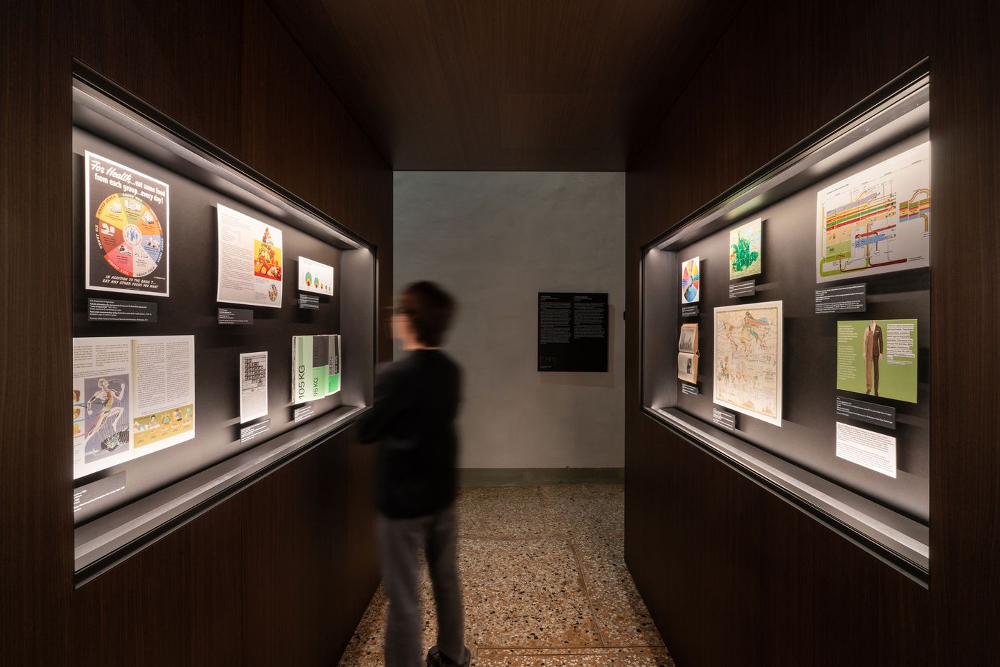 A dimly lit exhibition space with illuminated displays showcasing various posters, diagrams, and texts along the walls. A dimly lit exhibition space with illuminated displays showcasing various posters, diagrams, and texts along the walls.
