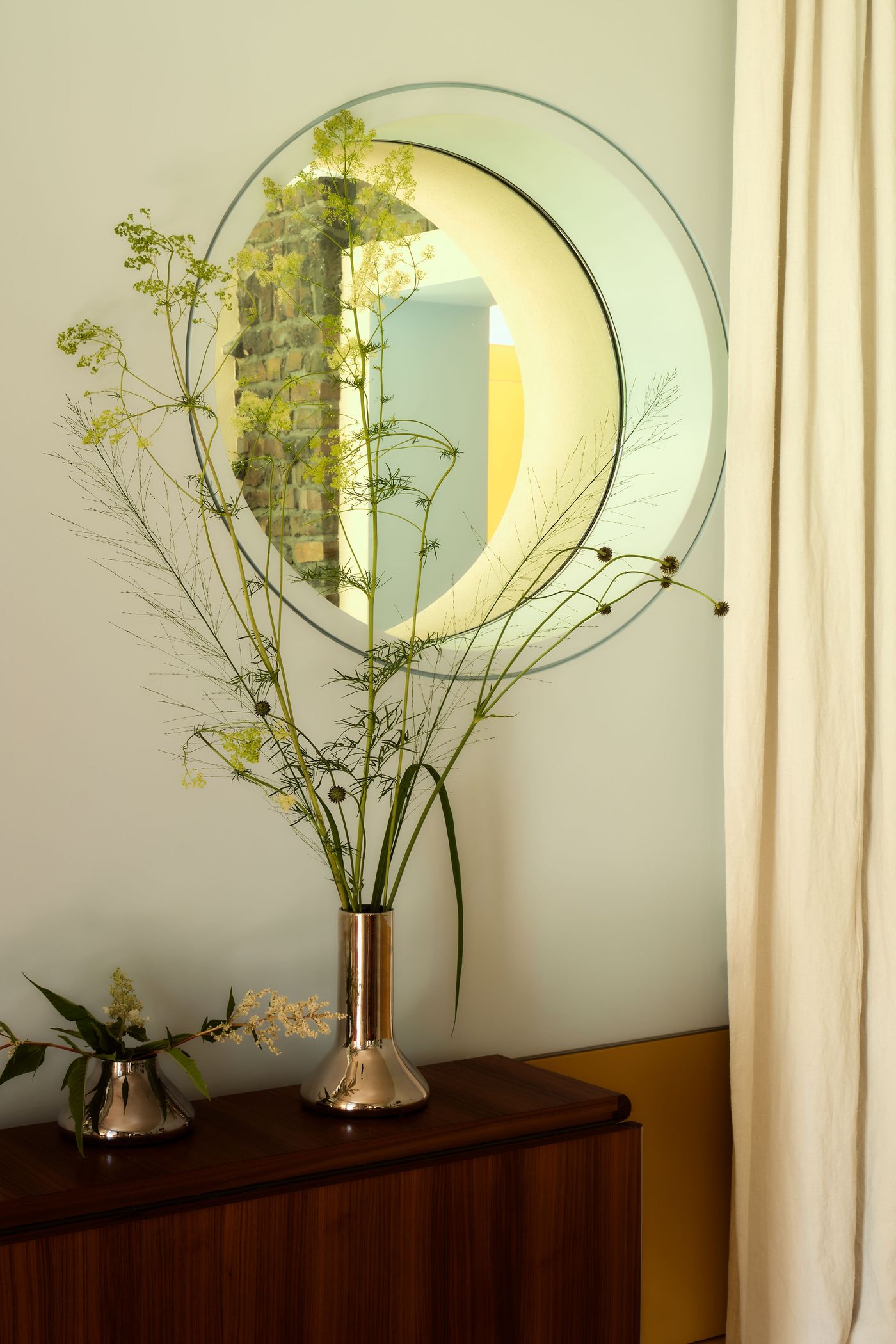 A modern interior featuring tall green flowering branches in a sleek vase, reflecting in a circular mirror beside a curtain.