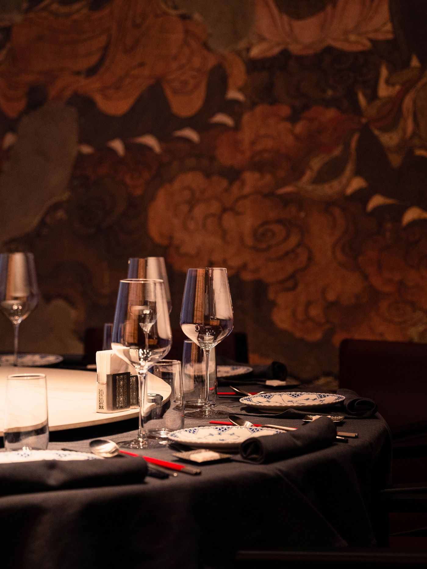 Elegant dining table set with glassware, plates, and utensils, against a richly patterned wall mural.