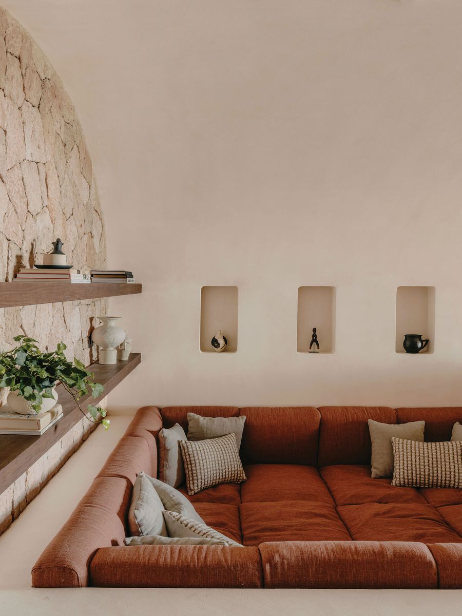 Cozy, rust-colored couch with plush cushions against a textured stone wall, featuring minimalist shelves and decorative items. Cozy, rust-colored couch with plush cushions against a textured stone wall, featuring minimalist shelves and decorative items.