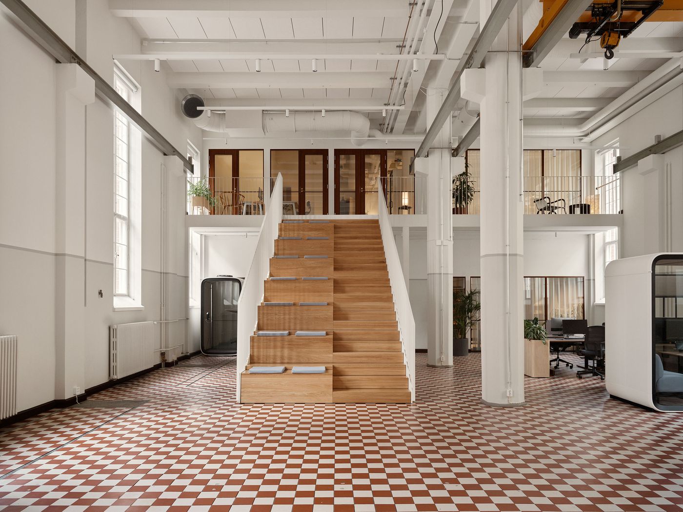 A grand wooden staircase with integrated shelving for books or decor dominates an open-plan office space, leading to an upper mezzanine. The vibrant red and white checkerboard floor complements the minimalist white walls and industrial accents, creating a dynamic and functional interior. A grand wooden staircase with integrated shelving for books or decor dominates an open-plan office space, leading to an upper mezzanine. The vibrant red and white checkerboard floor complements the minimalist white walls and industrial accents, creating a dynamic and functional interior.