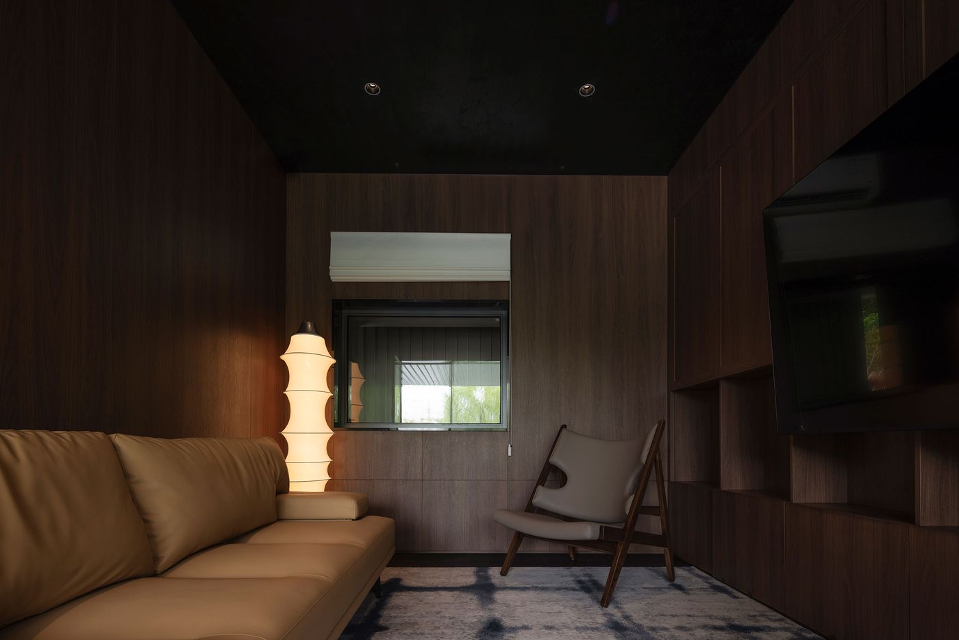 A cozy, darkened interior space in Wutopia Lab's Lake House is defined by dark wood paneling on the walls and integrated shelving, evoking a refined, moody atmosphere. A plush, light brown sofa and a minimalist canvas chair provide seating, while a unique sculptural floor lamp emits a warm, ambient light, subtly contrasting with the dark palette and enhancing the room's intimate and sophisticated character. A cozy, darkened interior space in Wutopia Lab's Lake House is defined by dark wood paneling on the walls and integrated shelving, evoking a refined, moody atmosphere. A plush, light brown sofa and a minimalist canvas chair provide seating, while a unique sculptural floor lamp emits a warm, ambient light, subtly contrasting with the dark palette and enhancing the room's intimate and sophisticated character.
