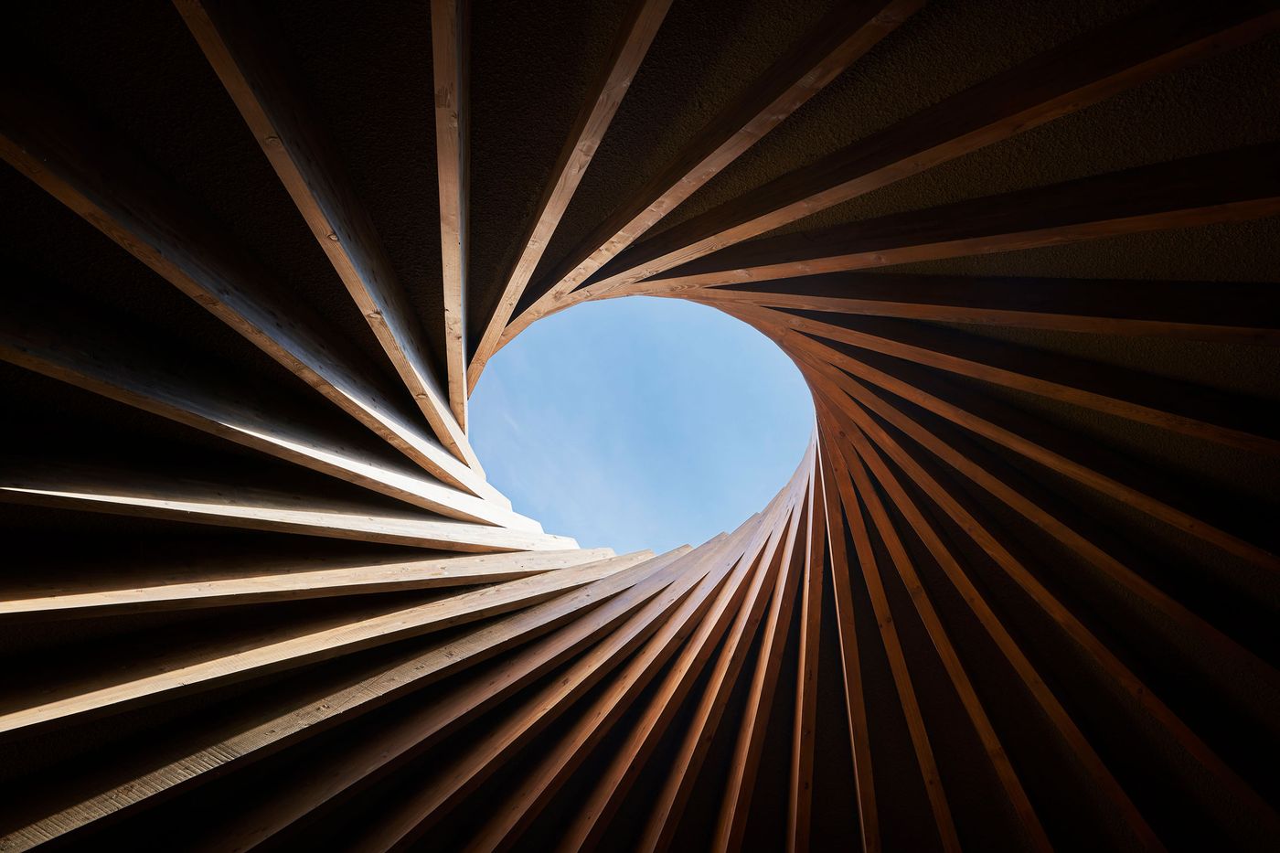 A spiral wooden structure opens towards a bright blue sky, creating a dynamic contrast between natural light and earthy tones. A spiral wooden structure opens towards a bright blue sky, creating a dynamic contrast between natural light and earthy tones.