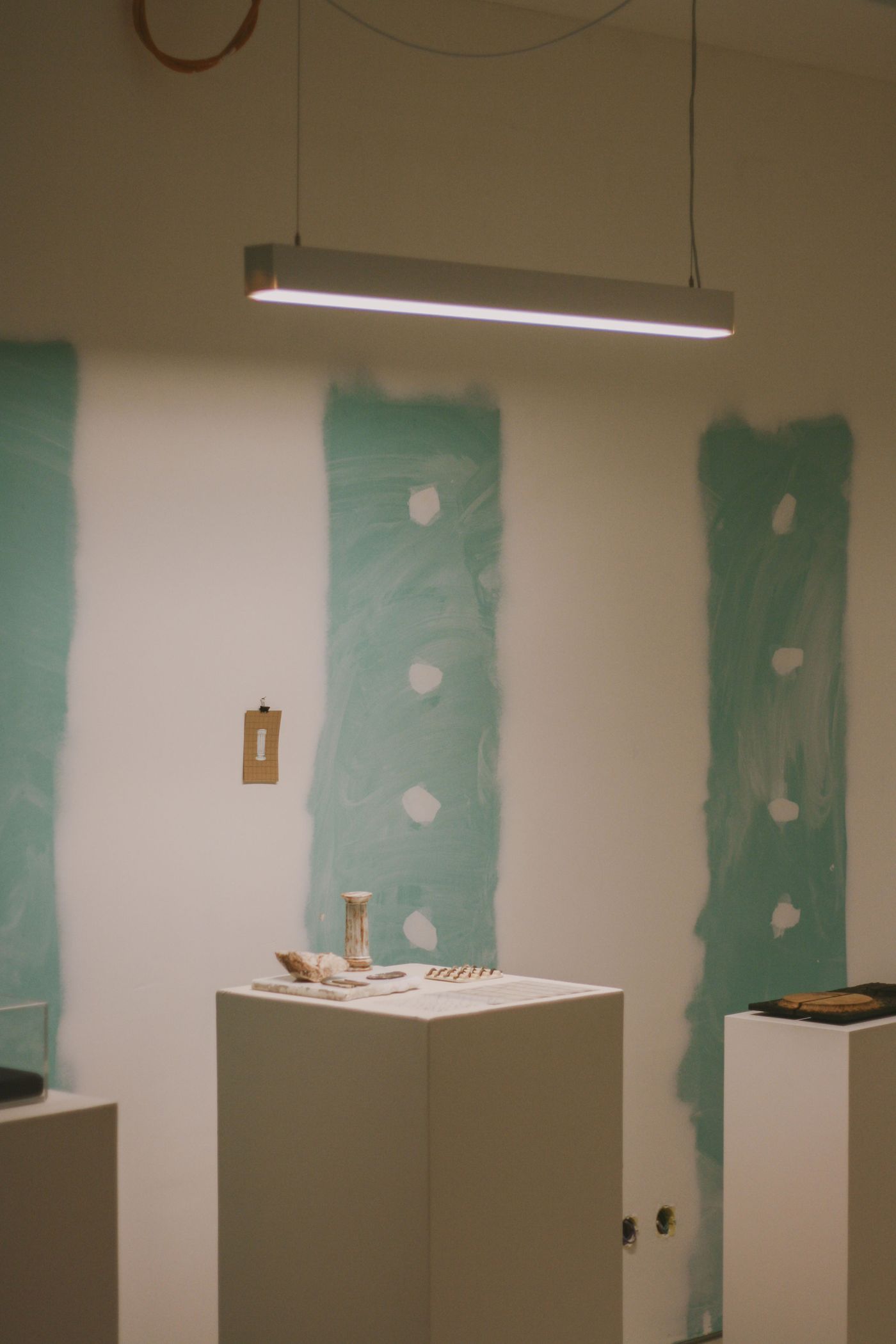 An exhibition space from "Crafting with AI" at Estrela 74 features a series of white pedestals displaying small, artisanal objects under a modern linear light fixture. The walls are partially painted with broad, raw teal strokes and visible plaster patches, creating a deconstructed industrial backdrop that contrasts with the refined display.