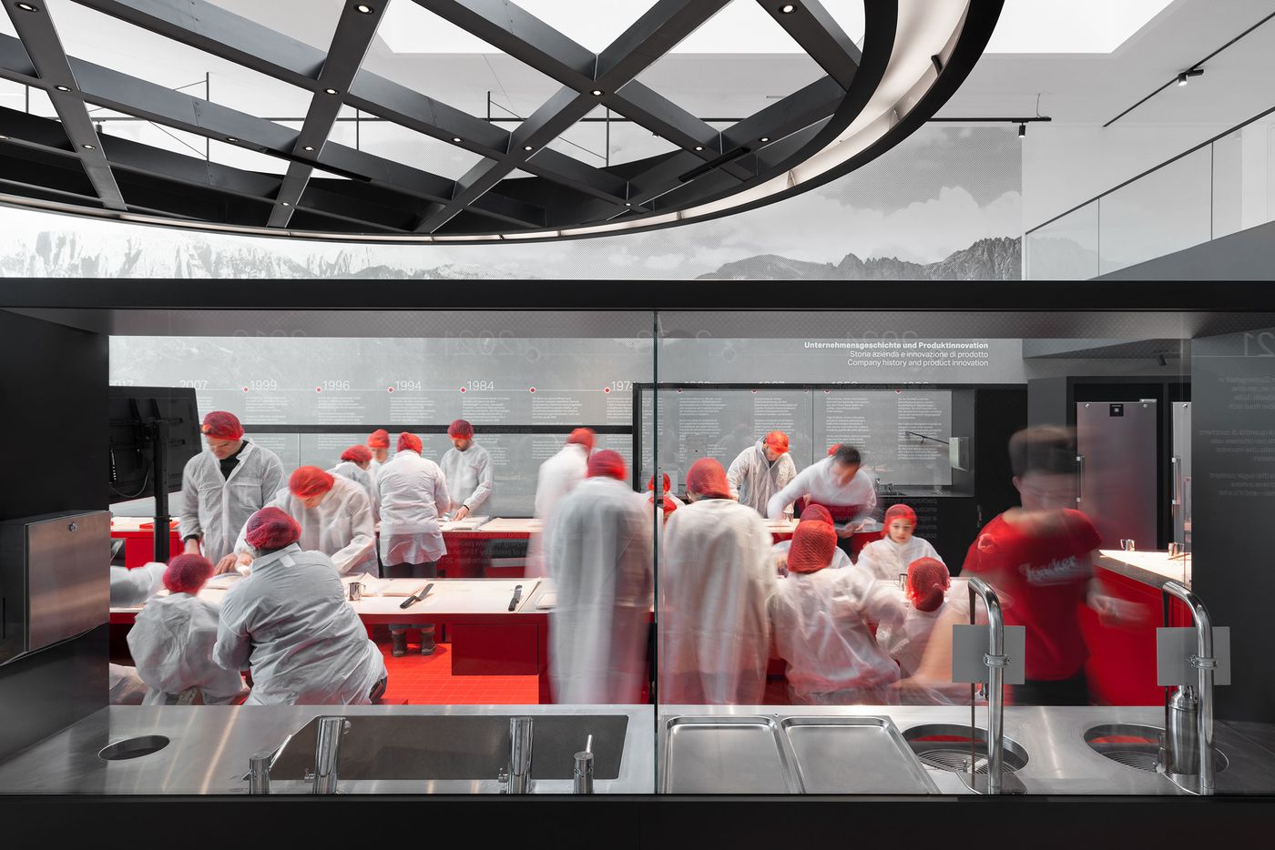 A live demonstration kitchen visible through floor-to-ceiling glazing transforms food production into theater. White-clad figures move through the red-accented workspace while visitors observe from sleek viewing stations, creating transparency between process and product that elevates manufacturing into cultural spectacle through architectural choreography. A live demonstration kitchen visible through floor-to-ceiling glazing transforms food production into theater. White-clad figures move through the red-accented workspace while visitors observe from sleek viewing stations, creating transparency between process and product that elevates manufacturing into cultural spectacle through architectural choreography.