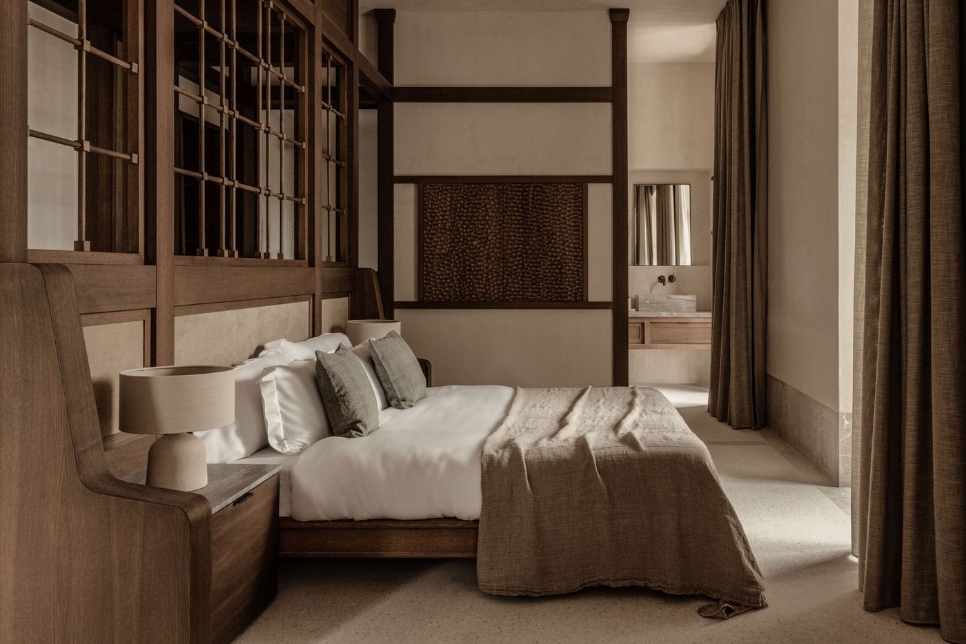 This elegant suite showcases a sophisticated Japandi design, with a prominent dark timber architectural frame separating the sleeping area from the bathroom. A large, textural woven art piece hangs on the wall. The bed, dressed in crisp white and soft grey linens, is flanked by modern table lamps, creating a warm, inviting, and meticulously crafted space. This elegant suite showcases a sophisticated Japandi design, with a prominent dark timber architectural frame separating the sleeping area from the bathroom. A large, textural woven art piece hangs on the wall. The bed, dressed in crisp white and soft grey linens, is flanked by modern table lamps, creating a warm, inviting, and meticulously crafted space.