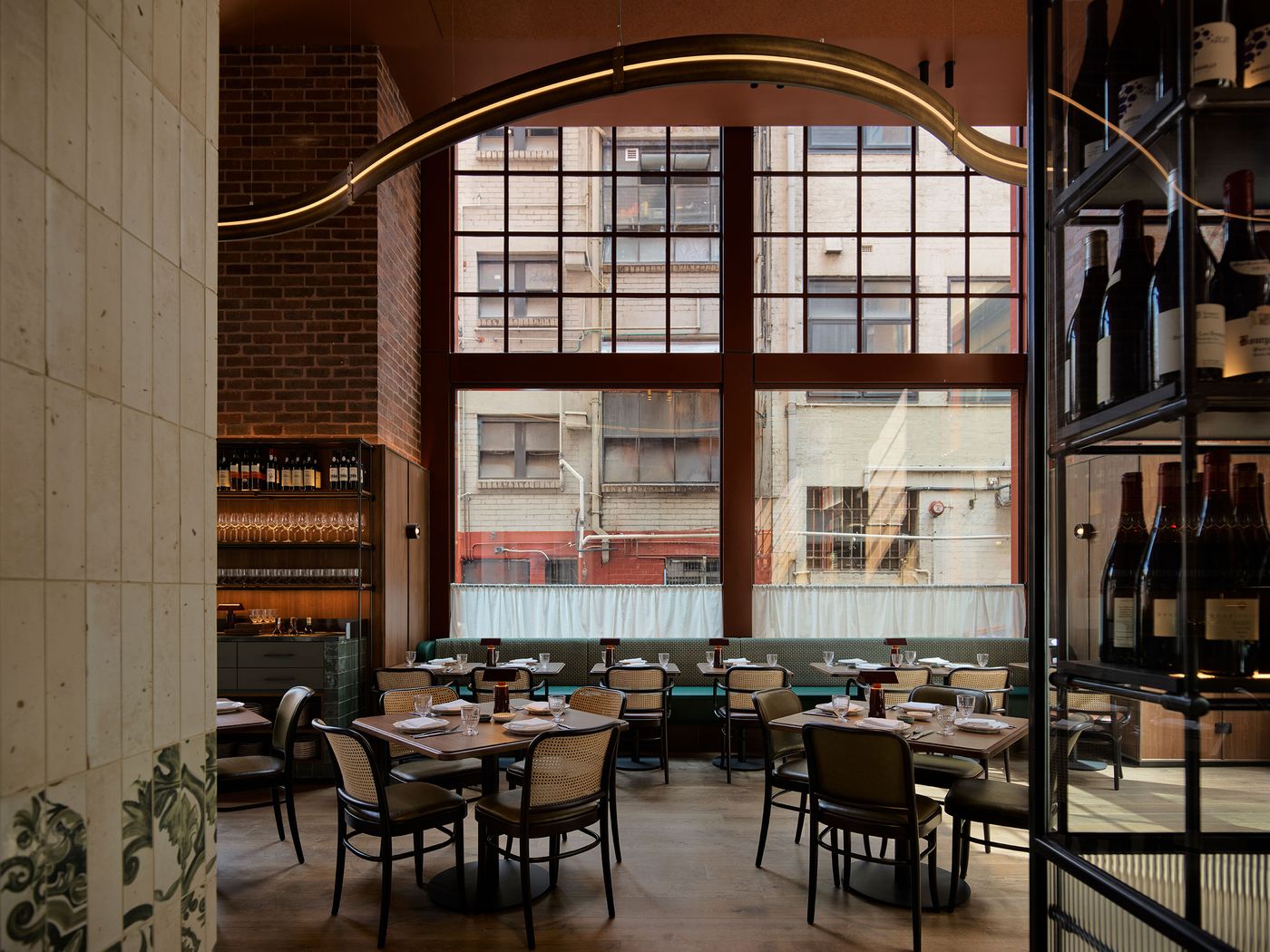 A modern restaurant interior featuring wooden tables, seating, and large windows showcasing an urban view, with wine bottles displayed. A modern restaurant interior featuring wooden tables, seating, and large windows showcasing an urban view, with wine bottles displayed.