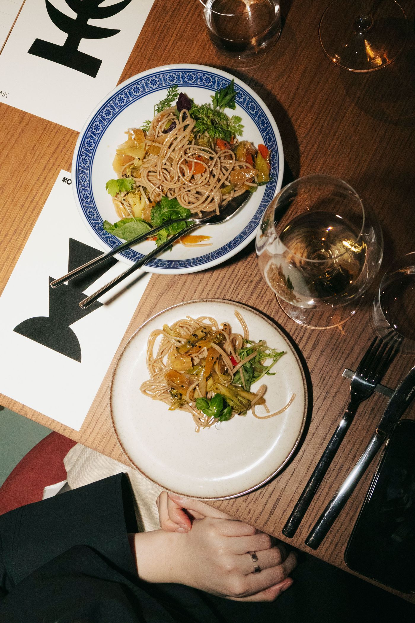Two plates of soba noodles with vegetables on a wooden table, accompanied by wine glasses and a menu. A person's hands are visible.