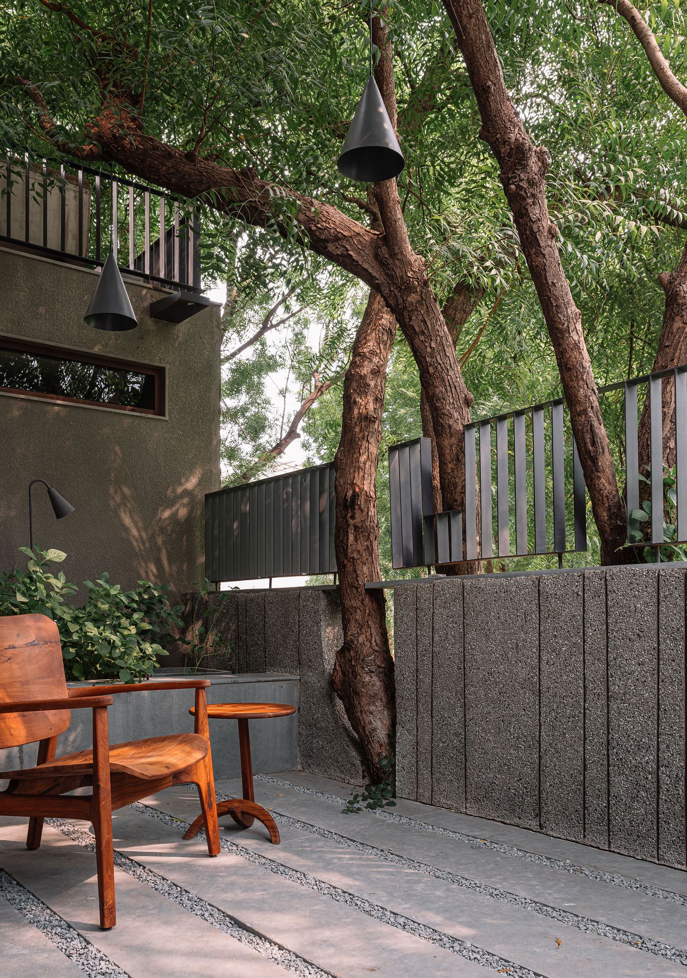An inviting outdoor patio, featuring a rustic wooden chair and a small side table, nestled amidst a grove of mature trees that punctuate the space. Industrial-style cone-shaped pendant lights hang from above, complementing the textured concrete walls and slatted metal fencing. The design harmonizes raw materials with lush biophilic elements, creating a tranquil retreat.