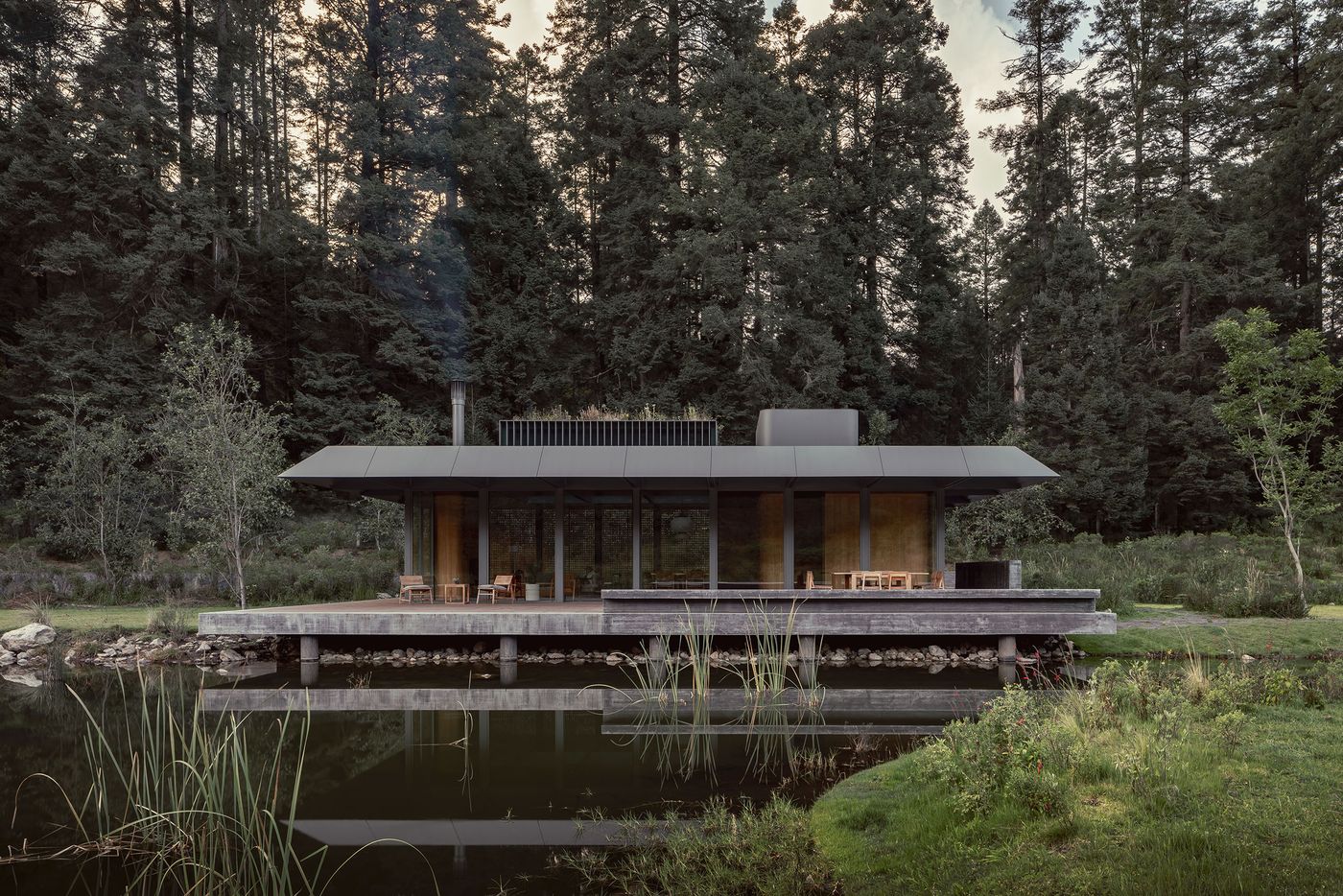 Modern wooden house by a tranquil pond, surrounded by tall trees, featuring a flat roof and large glass windows.