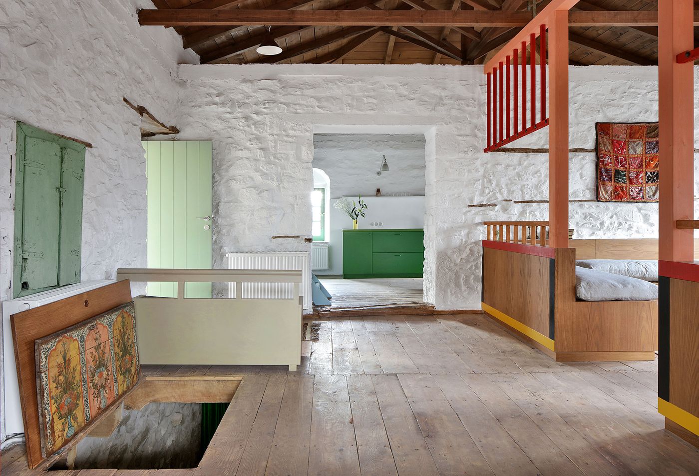 Wide-plank oak flooring grounds the multi-level interior, where sage-green built-ins complement the coral-red structural framework. Traditional elements like painted wood furniture and textiles create authentic Mediterranean character while contemporary spatial organization enables flexible community living arrangements.