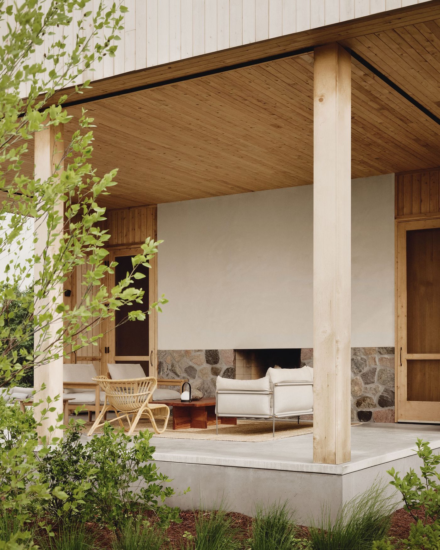 A modern porch with wooden beams and stone accents, featuring a white sofa, rattan chair, and lush greenery.
