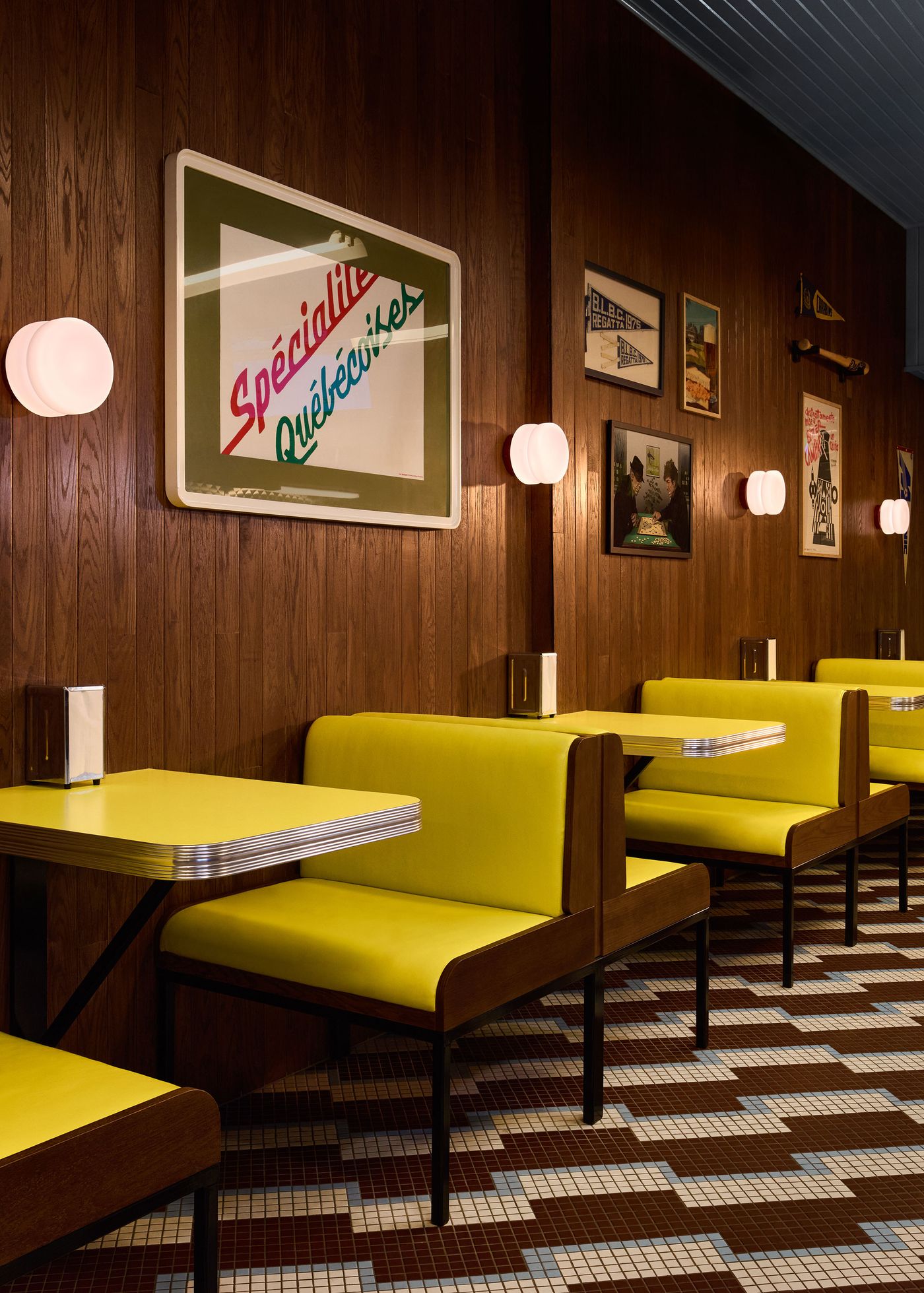 The interior of Renzo Sandwich is captured, showcasing a row of bright yellow diner booths against warm wood-paneled walls. The space is illuminated by soft, modern globe lights. A prominent sign reads "Spécialité Québécoise," and the distinctive geometric pattern of the tiled floor completes the retro-diner aesthetic.