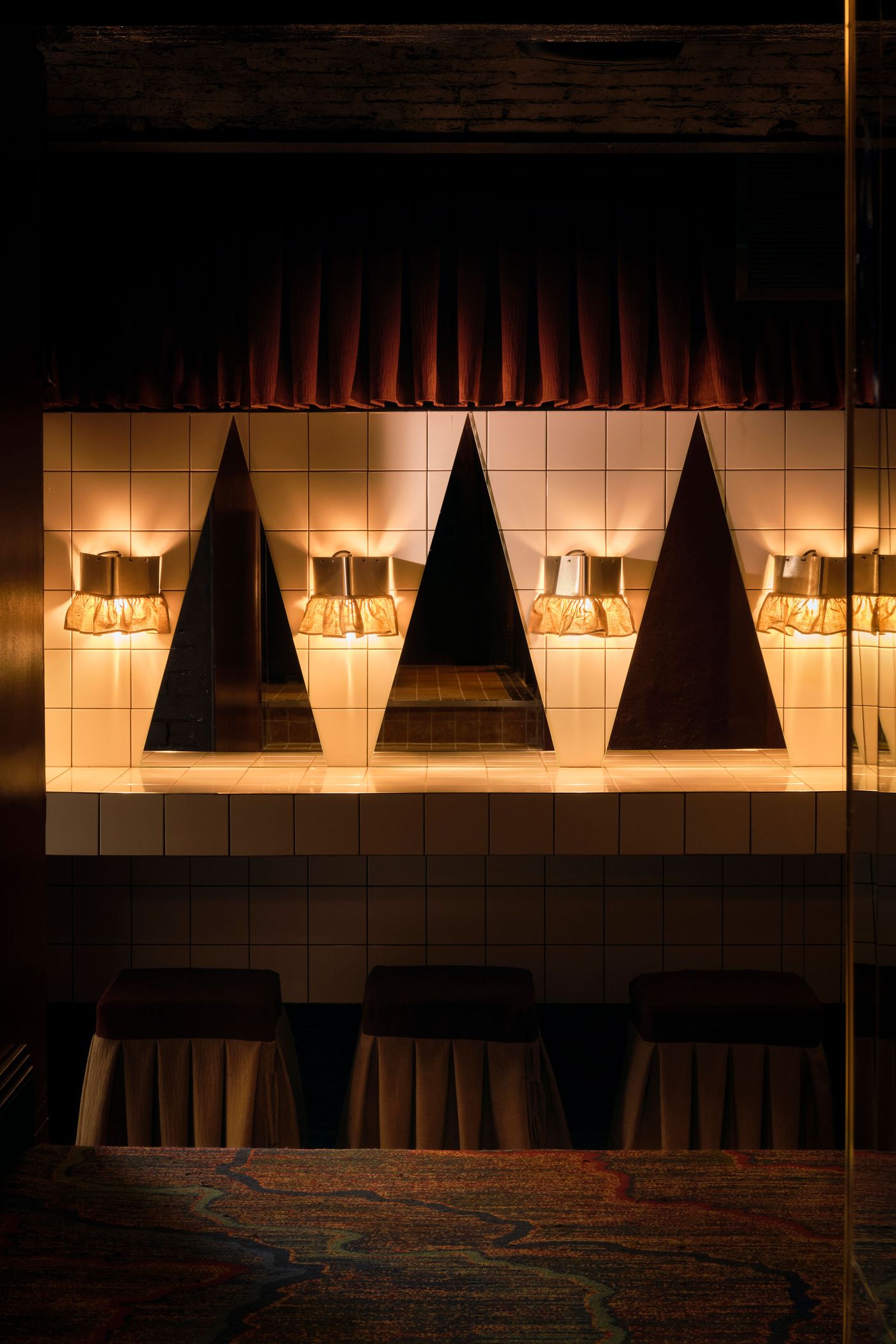 A unique and visually striking wall installation in The Roses of Elagabalus features a series of triangular mirrored panels set into a tiled wall. Between these panels, vintage sconces with ruffled shades emit a warm, inviting light. Below, plush, skirted stools sit on a richly patterned carpet, creating an eclectic and playful yet sophisticated space. A unique and visually striking wall installation in The Roses of Elagabalus features a series of triangular mirrored panels set into a tiled wall. Between these panels, vintage sconces with ruffled shades emit a warm, inviting light. Below, plush, skirted stools sit on a richly patterned carpet, creating an eclectic and playful yet sophisticated space.