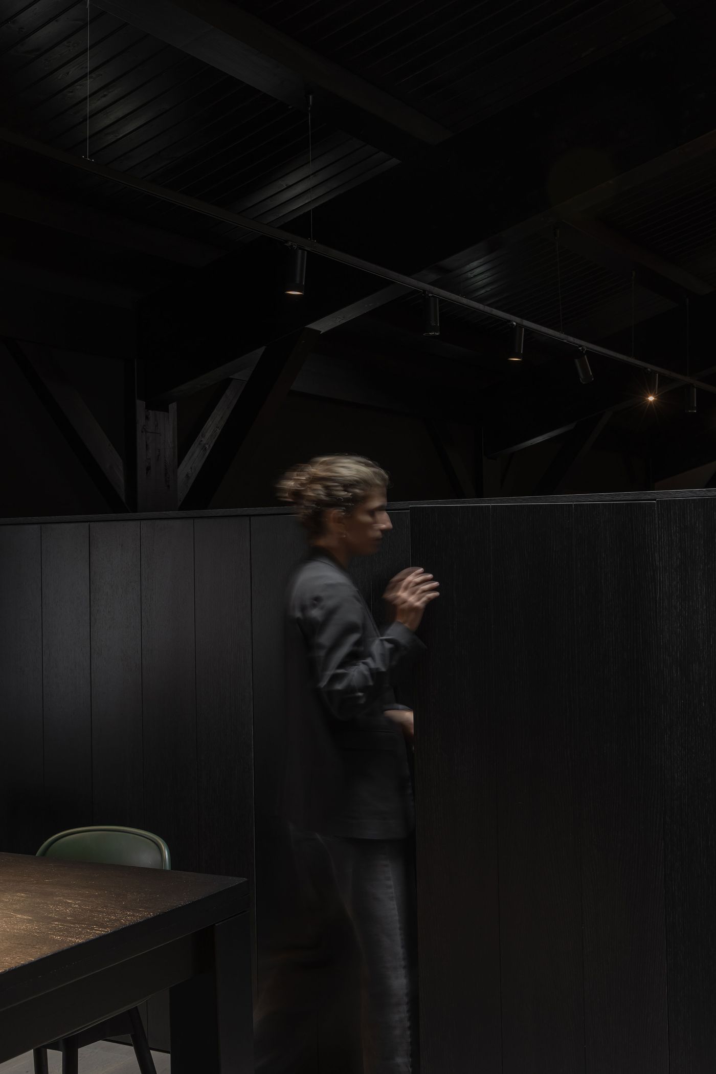 A person in dark clothing walks past tall black partitions in a dimly lit interior space with wooden beams and a simple table.