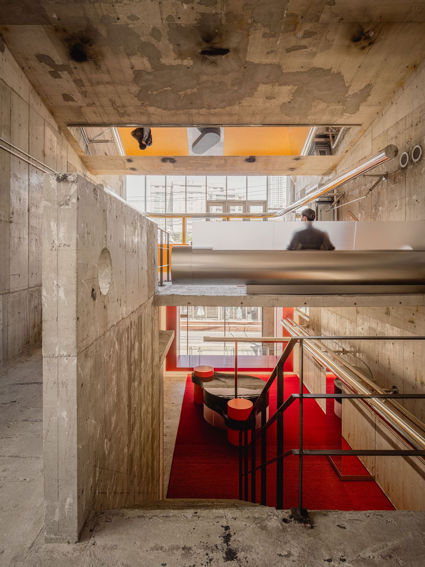 A sectional view through ANDERSSON BELL Tokyo reveals the store’s layered circulation, with rough concrete slabs framing glimpses of the yellow upper floor and red ground level below. A visitor occupies the intermediate platform, while chrome rails, black balustrades and sculptural displays reinforce I IN’s interplay of openness, colour and industrial structure.