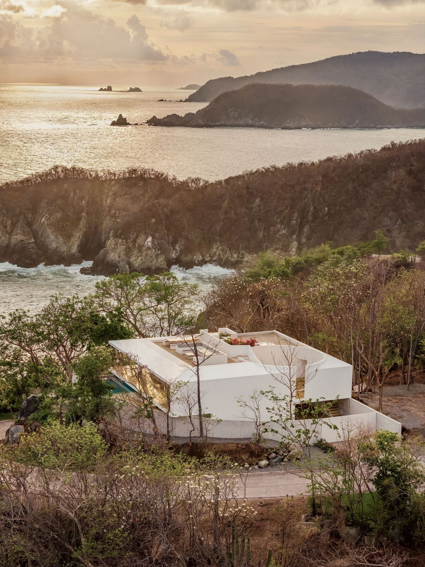Low golden light washes over Casa Malonic and the coastline beyond, where layered headlands and scattered sea stacks fade into a glinting Pacific. Closer in, the house's cantilevered white roof and circular courtyard emerge from dry tropical forest, its glazed living level catching the sun above a sliver of turquoise pool. Low golden light washes over Casa Malonic and the coastline beyond, where layered headlands and scattered sea stacks fade into a glinting Pacific. Closer in, the house's cantilevered white roof and circular courtyard emerge from dry tropical forest, its glazed living level catching the sun above a sliver of turquoise pool.