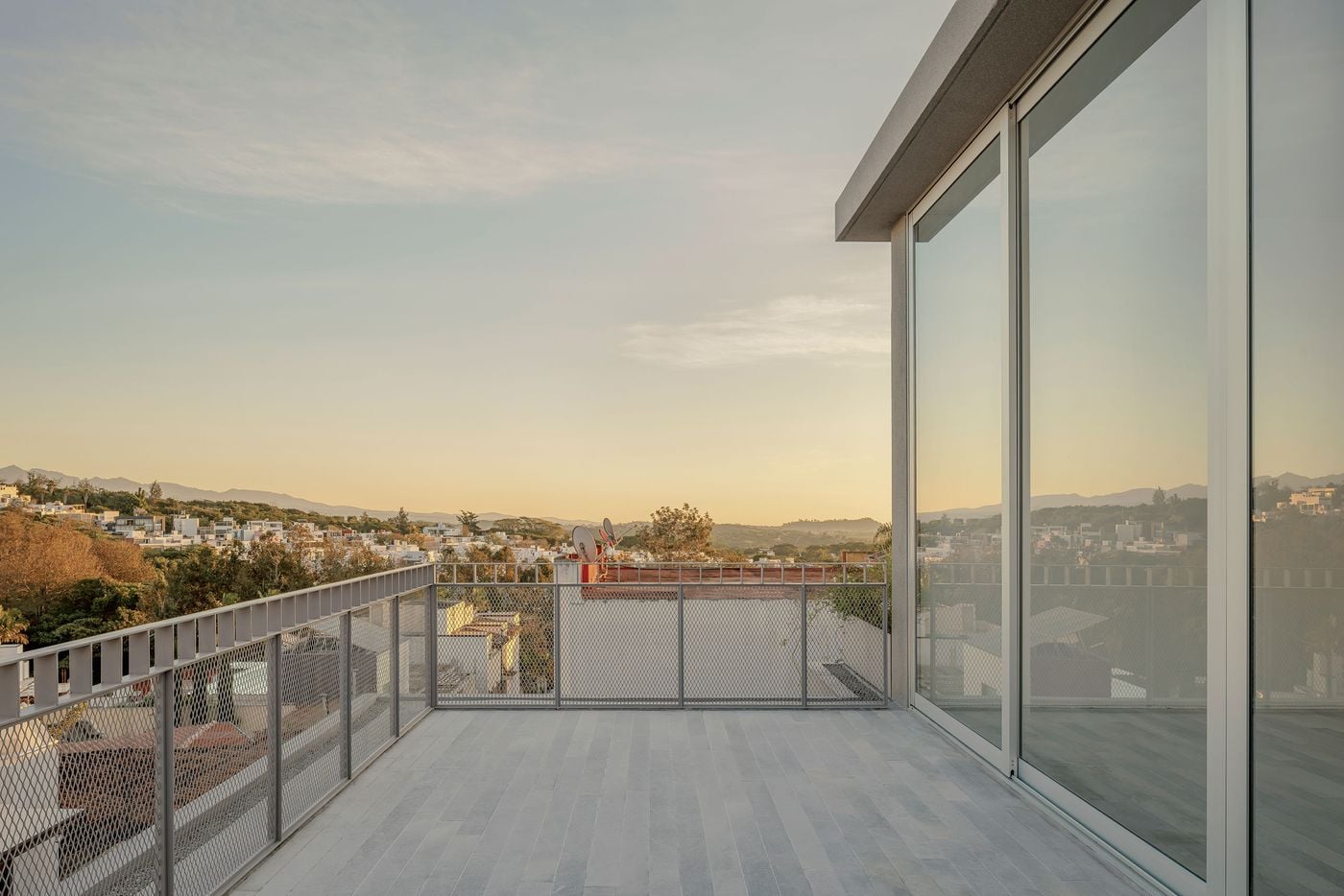 Casa Tlaloc’s upper terrace opens broadly towards Xalapa’s low-rise neighbourhood and distant mountain ridges. Pale grey paving and utilitarian metal-mesh balustrades maintain the house’s restrained palette, while expansive glazing reflects the evening landscape, allowing architecture and horizon to merge in the warm light of sunset. Casa Tlaloc’s upper terrace opens broadly towards Xalapa’s low-rise neighbourhood and distant mountain ridges. Pale grey paving and utilitarian metal-mesh balustrades maintain the house’s restrained palette, while expansive glazing reflects the evening landscape, allowing architecture and horizon to merge in the warm light of sunset.