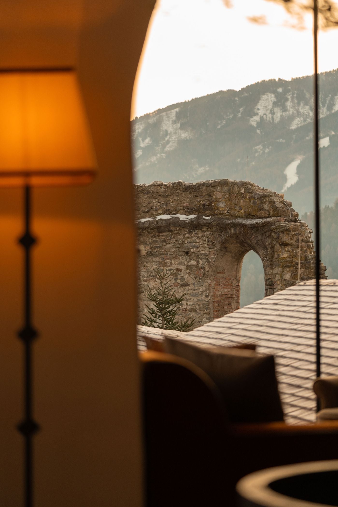 Seen from a lamplit interior at Castel Badia, a tall arched window frames the weathered ruins of a medieval stone wall pierced by a single arch. A shingled roof lies in the foreground and forested, snow-streaked slopes rise beyond, while a glowing floor lamp and silhouetted armchair warm the foreground. Seen from a lamplit interior at Castel Badia, a tall arched window frames the weathered ruins of a medieval stone wall pierced by a single arch. A shingled roof lies in the foreground and forested, snow-streaked slopes rise beyond, while a glowing floor lamp and silhouetted armchair warm the foreground.