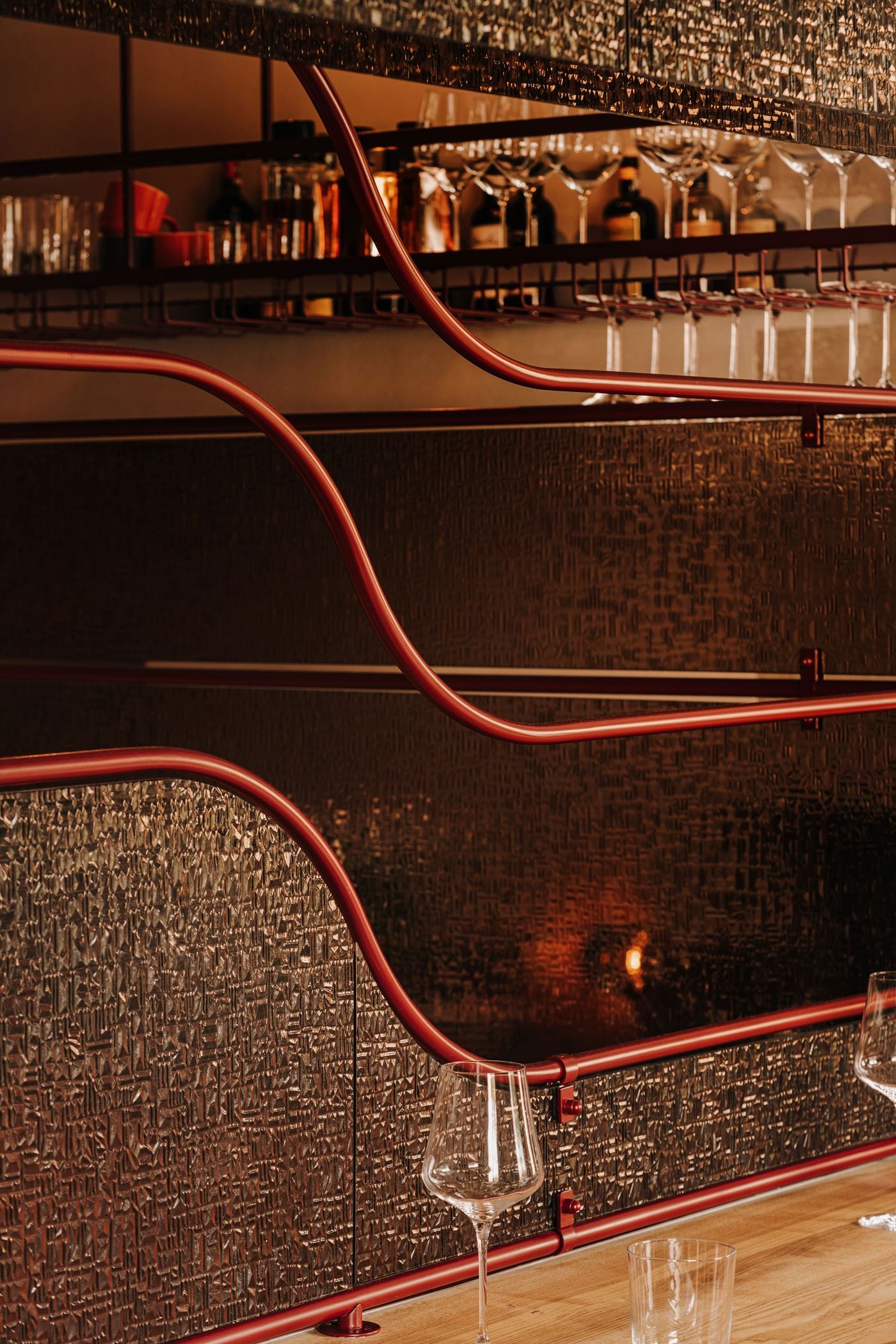 A close-up of the upstairs dining counter at WIN wine bar reveals sinuous burgundy metal rails framing richly textured glass partitions. Behind them, suspended racks hold rows of inverted wine glasses, while the warm lighting catches the irregular glass surfaces, creating shimmering reflections that contrast with the precision of the metalwork. A close-up of the upstairs dining counter at WIN wine bar reveals sinuous burgundy metal rails framing richly textured glass partitions. Behind them, suspended racks hold rows of inverted wine glasses, while the warm lighting catches the irregular glass surfaces, creating shimmering reflections that contrast with the precision of the metalwork.