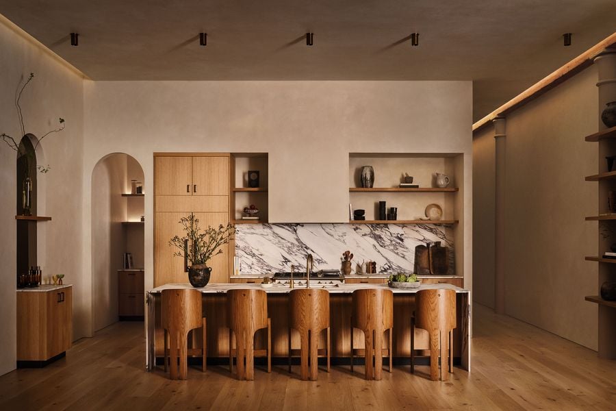 A modern kitchen featuring wooden cabinetry, a marble backsplash, and elegant wooden stools, accented by minimalist decor and soft lighting.