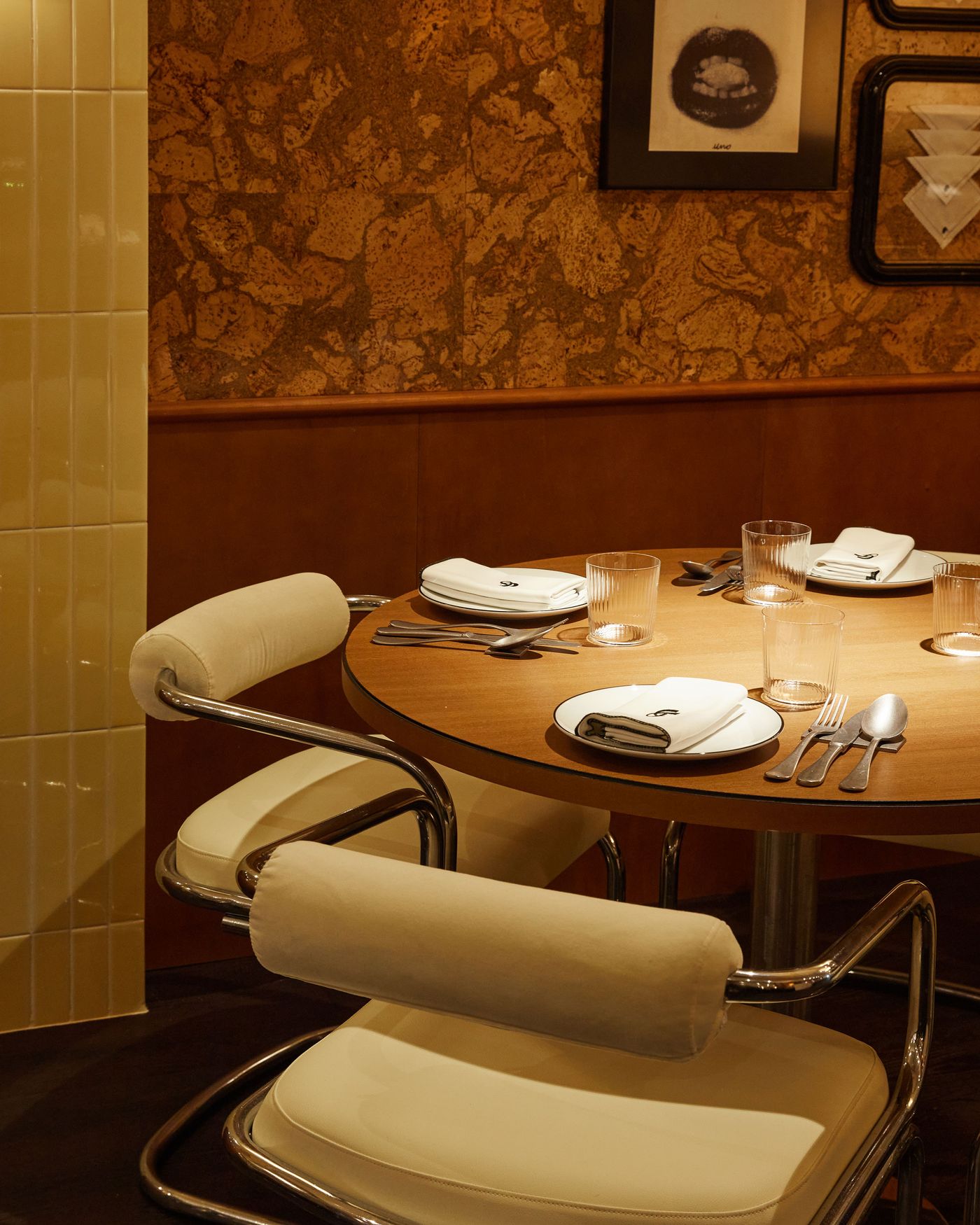 A close-up of a circular dining table set with minimalist white plates and glassware. The elegant mid-century modern chairs feature polished chrome frames and plush, cream-colored cushions. The backdrop is a textured wall with a warm, speckled pattern, creating a rich contrast between materials.