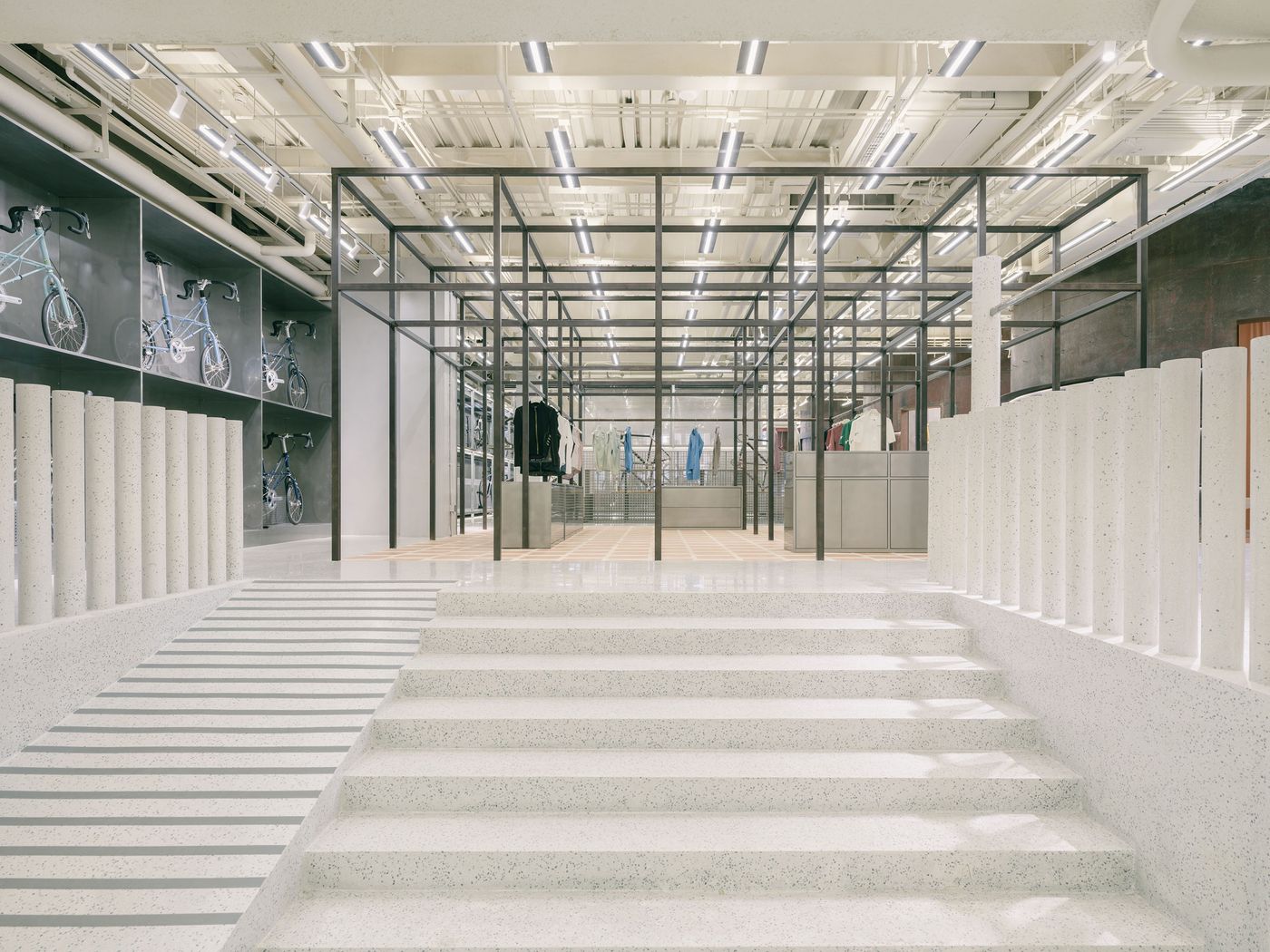 The entrance to AITASHOP is defined by a grand, minimalist staircase of light grey terrazzo, flanked by a parallel ramp with subtle linear grooves, and a striking balustrade of vertical white cylindrical posts. Beyond, a bold black metal grid structure forms the core retail area, showcasing bicycles against a backdrop of varied textures and modern lighting. The entrance to AITASHOP is defined by a grand, minimalist staircase of light grey terrazzo, flanked by a parallel ramp with subtle linear grooves, and a striking balustrade of vertical white cylindrical posts. Beyond, a bold black metal grid structure forms the core retail area, showcasing bicycles against a backdrop of varied textures and modern lighting.