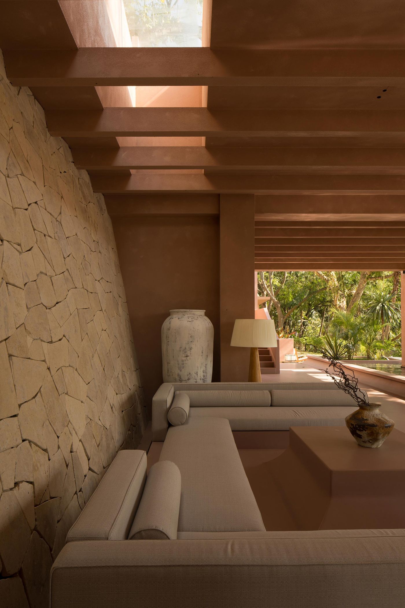 A cozy, earthy-toned living space featuring stone walls, modern sofas, a decorative vase, and a view of lush greenery through large windows. A cozy, earthy-toned living space featuring stone walls, modern sofas, a decorative vase, and a view of lush greenery through large windows.