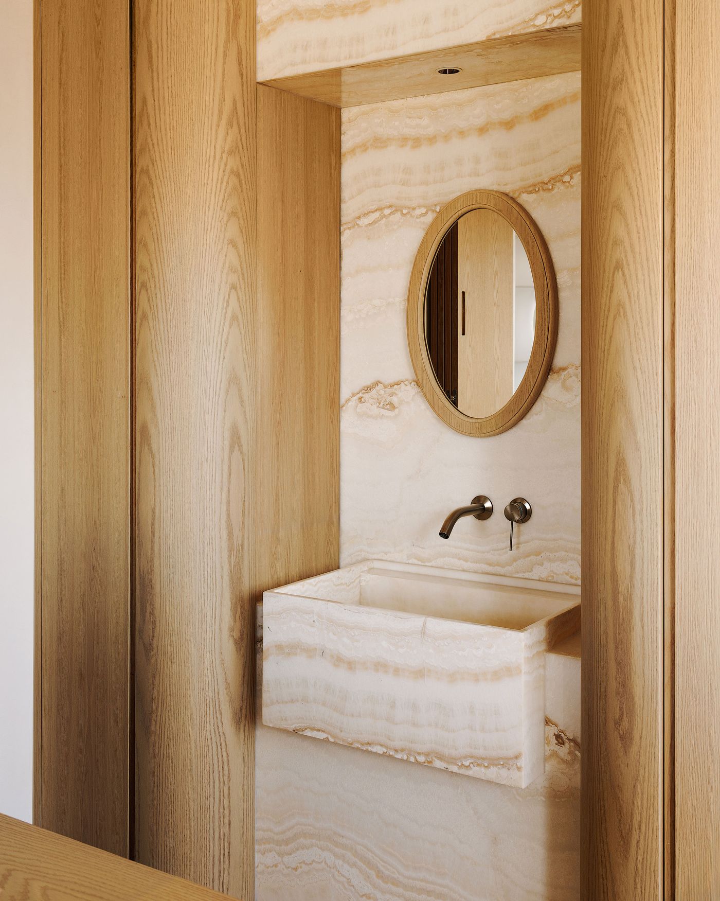 A luxurious and artfully designed bathroom vanity area within a Shanghai apartment. The space features an elegant oval mirror set against a stunning veined onyx wall, with a matching contemporary onyx basin and minimalist bronze fixtures. Warm wooden paneling frames the alcove, creating a harmonious blend of natural materials and sophisticated design.