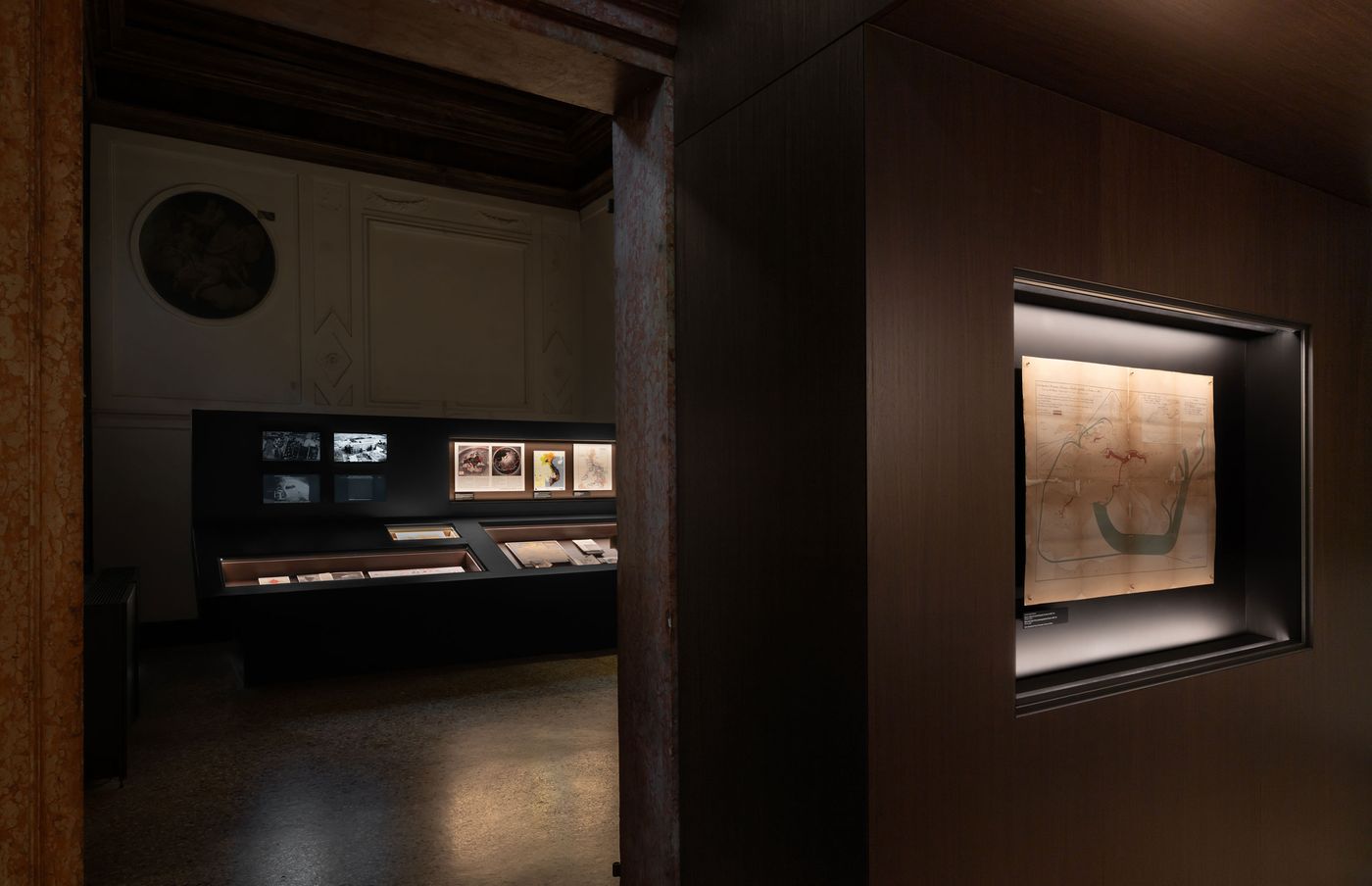 A dimly lit gallery exhibits historical documents and artwork, with a focus on a framed, detailed map in a shadowed alcove. A dimly lit gallery exhibits historical documents and artwork, with a focus on a framed, detailed map in a shadowed alcove.