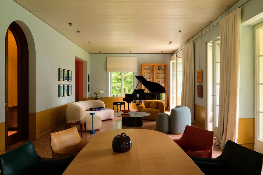 A stylish living room featuring a grand piano, colorful seating, and large windows, creating a bright and inviting atmosphere.