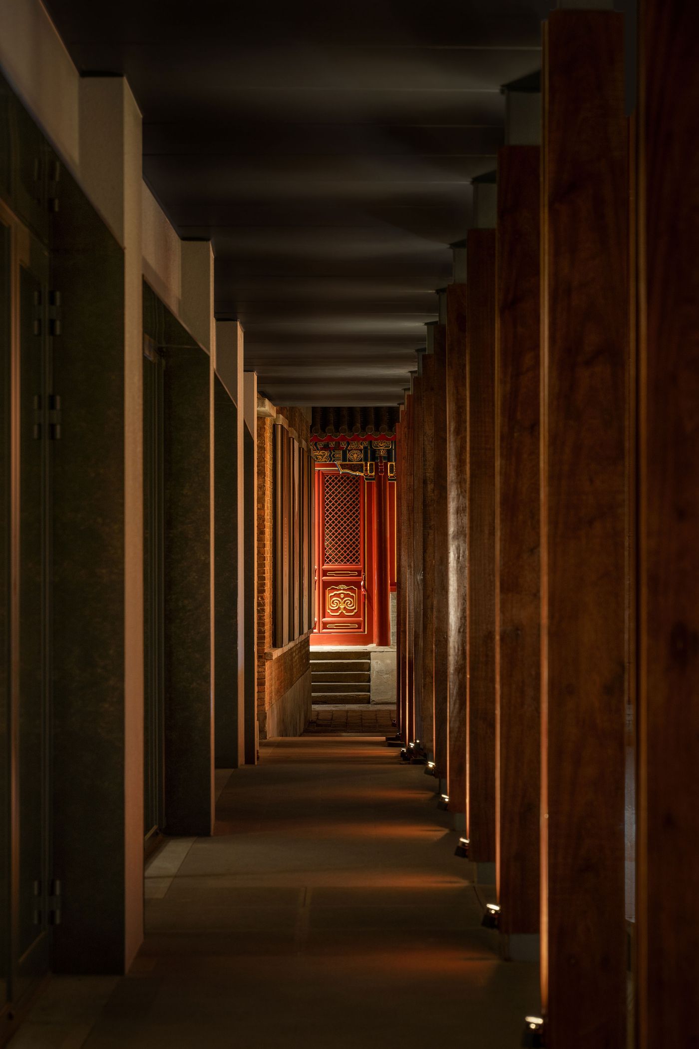 A view down a dimly lit corridor lined with wooden pillars, leading to a vibrant red door with ornate details and steps beyond. A view down a dimly lit corridor lined with wooden pillars, leading to a vibrant red door with ornate details and steps beyond.